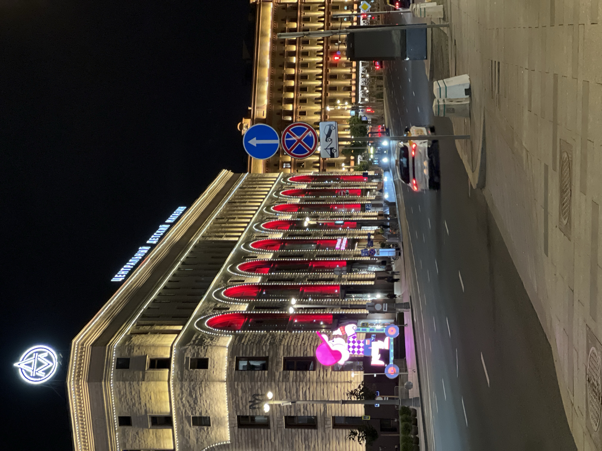 This is a night-time, eye-level, vertical shot of a wide city street in Moscow, Russia, in front of a large, illuminated building. The building on the left is a grand structure with classical architectural elements, featuring a prominent neon sign at its peak that reads "ЦЕНТРАЛЬНЫЙ ДЕТСКИЙ МАГАЗИН" (Central Children's Store) in Cyrillic letters. The facade is lit with warm yellow lights, and a series of arched windows or openings are highlighted with bright red lighting and white string lights. In front of this building, a large, colorful, illuminated sculpture of a cartoon character on a scooter stands on the sidewalk.
The street is a multi-lane asphalt road with clear white lane markings. Several cars are visible, including a white car in the foreground on the right lane and another white car in the distance on the left lane. Traffic lights are visible, appearing red in the distance.
On the right side of the street, a sidewalk is paved with stone tiles. Several traffic signs are mounted on poles on the sidewalk, including a blue circular sign with a white arrow pointing up, a red and blue circular sign with a red cross, and a white rectangular sign depicting a tow truck. The sidewalk is lined with bollards.
In the background, another large building is visible, also illuminated with warm yellow lights. The overall atmosphere is that of a bustling urban environment at night, with prominent lighting and commercial signage.