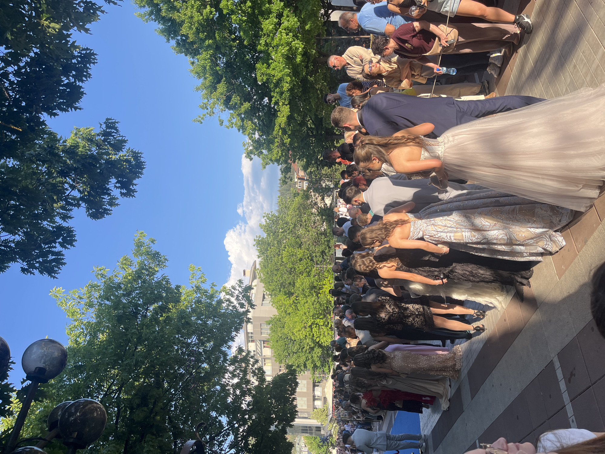 The image shows a crowd gathered along a brick walkway under trees in Blagoevgrad, Bulgaria. The sky is blue with some clouds. The crowd includes people in formal attire, with many women in long dresses. Spectators are also present, watching the procession. Buildings are visible in the background.
