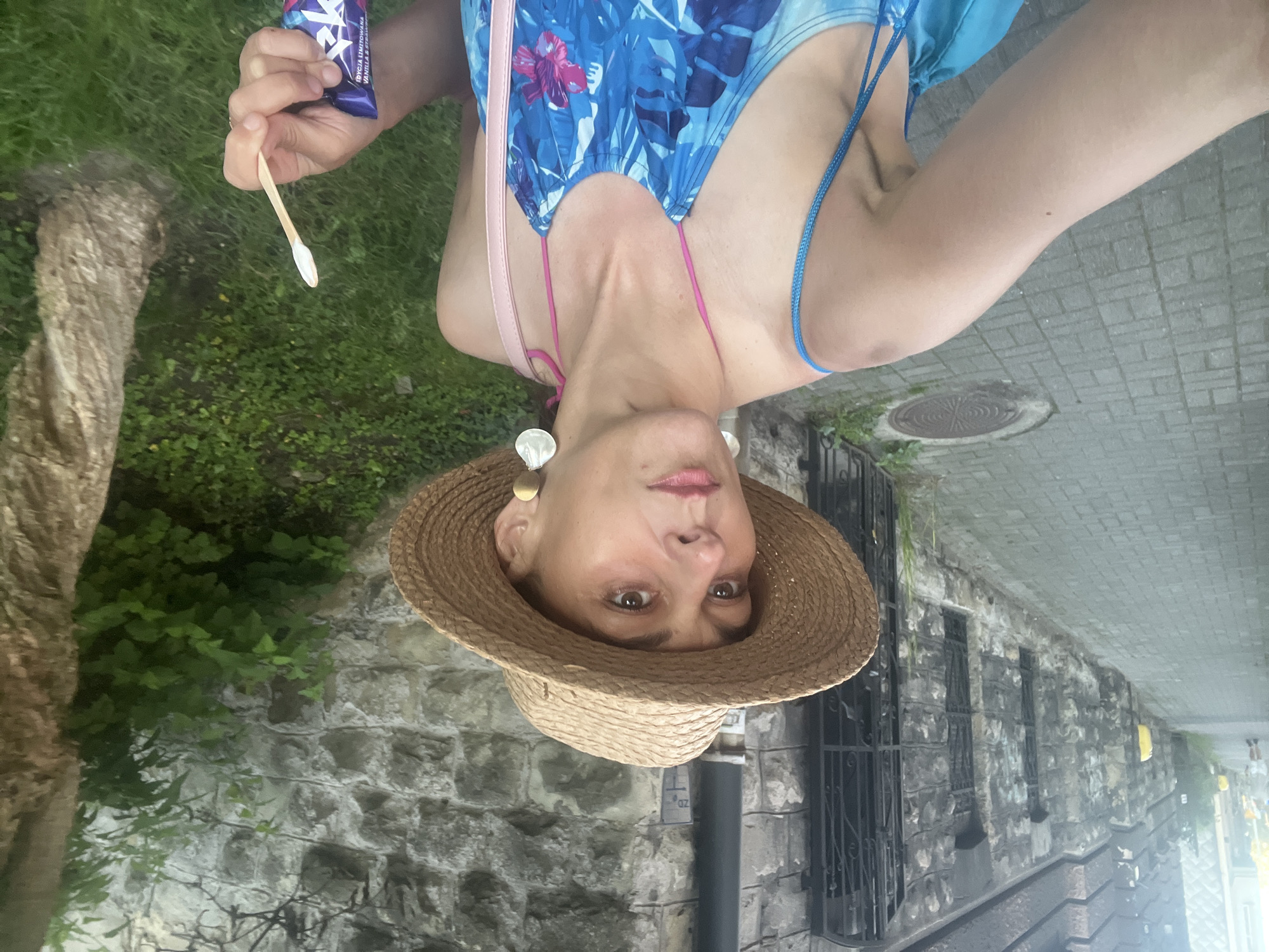 A woman in a straw hat and a blue floral dress stands on a paved sidewalk next to a stone building and a tree. She is holding a wooden popsicle stick with a white substance on it and a purple wrapper in her right hand. She is looking to her left. The scene is outdoors in an urban setting.