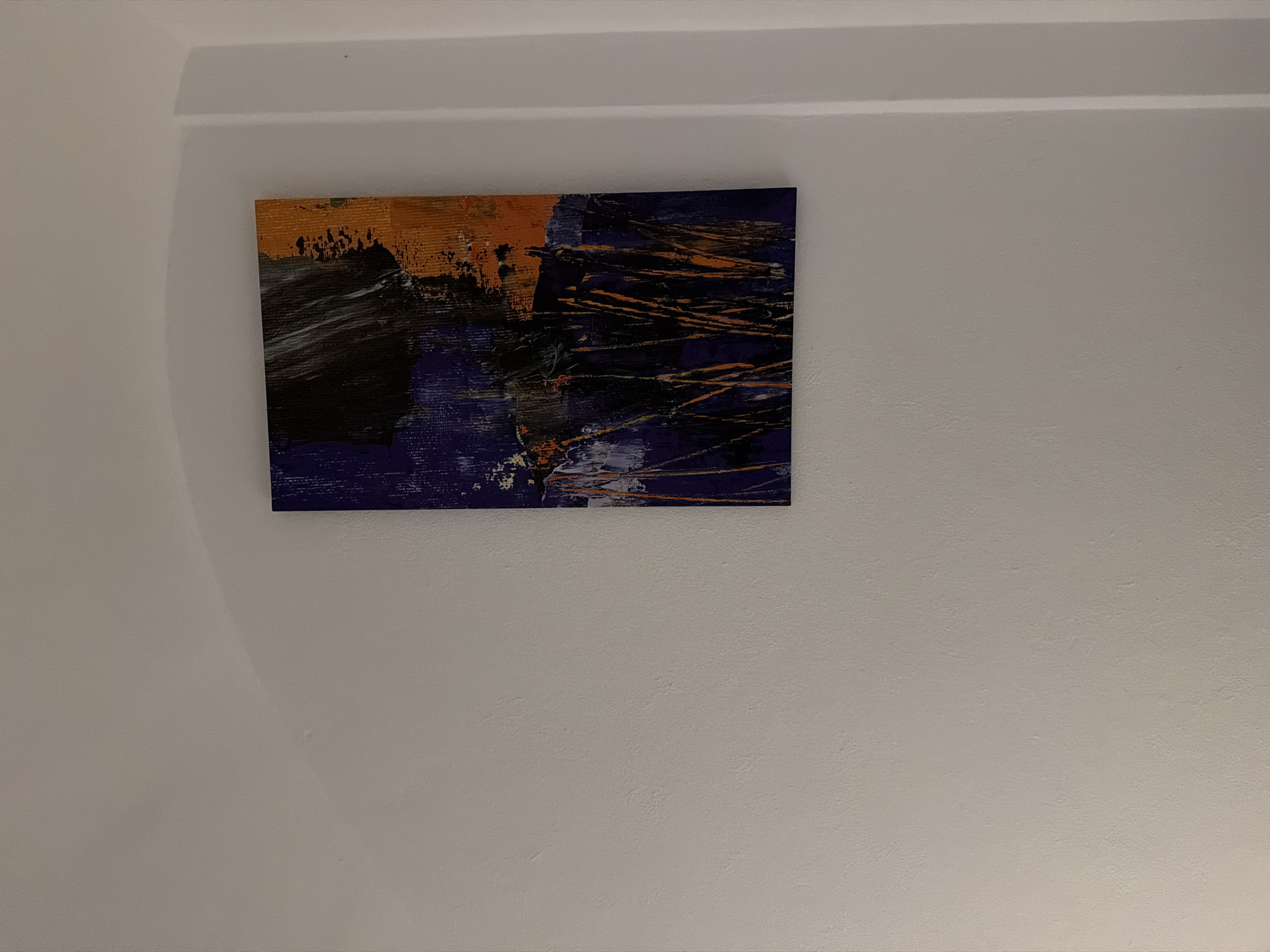 The media file shows a framed abstract painting hanging on a textured, off-white wall. The painting is square and features bold, energetic brushstrokes in shades of orange, black, and deep blue, with some white accents. The orange appears in a textured block at the top left, contrasting with dark, sweeping strokes below it. The bottom portion of the painting is dominated by deep blue, interspersed with thin, irregular lines of orange and white, suggesting movement or a chaotic energy. The style is modern and expressive, with visible texture from the paint application.
The wall on which the painting hangs has a subtle texture, characteristic of interior walls. Above the painting, a white architectural molding runs along the ceiling line, creating a shadow that emphasizes the curve of the wall and ceiling junction. The lighting is soft, casting faint shadows and highlighting the textured surfaces.
The scene appears to be an interior setting, possibly a living room, hallway, or gallery space. There are no people or discernible objects other than the painting and the wall. The overall impression is one of contemporary art displayed in a simple, uncluttered environment. The time of day and weather are not discernible. No text is visible in the media file. Given the location context of Sant Antoni De Portmany, Spain, this could be a piece of art in a home or a commercial establishment on the island.