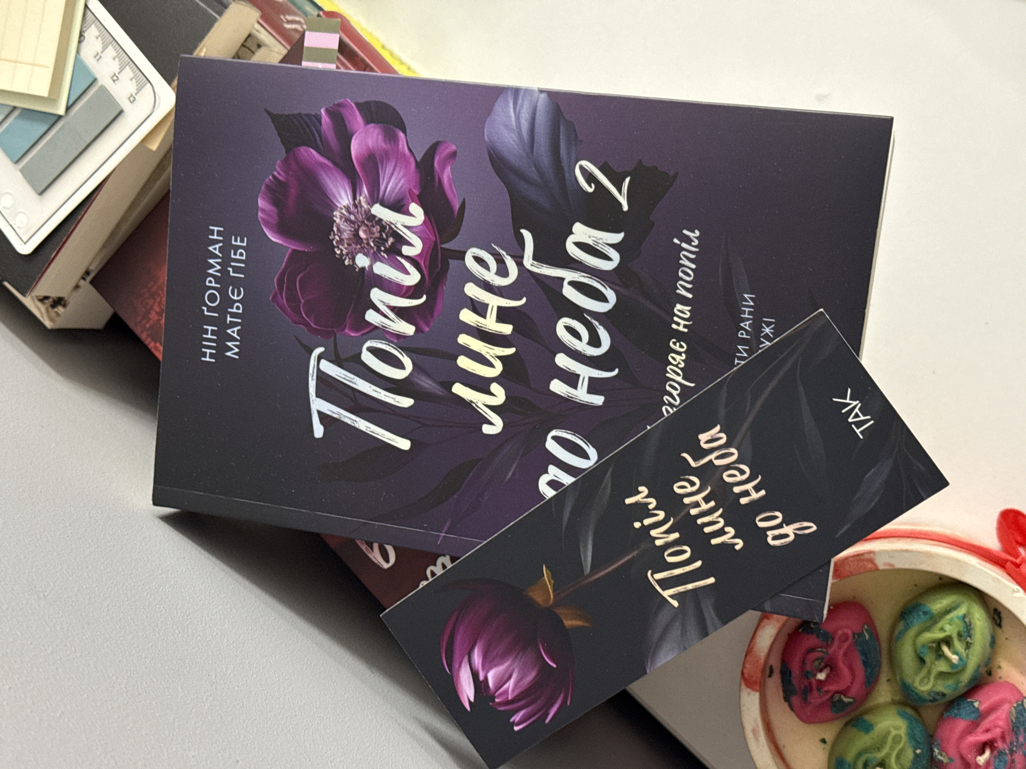 This media file shows a close-up view of a book lying on a surface, possibly a table or desk. The book has a dark purple cover featuring a large, stylized purple flower and the title "Попіл лине до неба 2" (Popil lyne do neba 2) in a stylized white font. Beneath the title, there is additional text that reads "НІН ҐОРМАН МАТЬЄ ҐІБЕ" and "горяє на попіл" along with "ТИ РАНИ УЖІ". A bookmark, also with a dark cover and a floral design, is placed over the book. This bookmark has the text "Попіл лине до неба" and "TAK".
The book and bookmark are positioned on a light-colored, textured surface, casting a soft shadow. In the background, other books and office supplies, such as sticky notes and a ruler, are partially visible, suggesting a workspace or study area. To the right, a small decorative item containing several colorful, rose-shaped candles is visible, adding a touch of decorative detail to the scene. The overall lighting suggests an indoor setting, likely during the daytime, with no specific weather conditions discernible. The arrangement implies a moment of pause, perhaps during reading or studying.