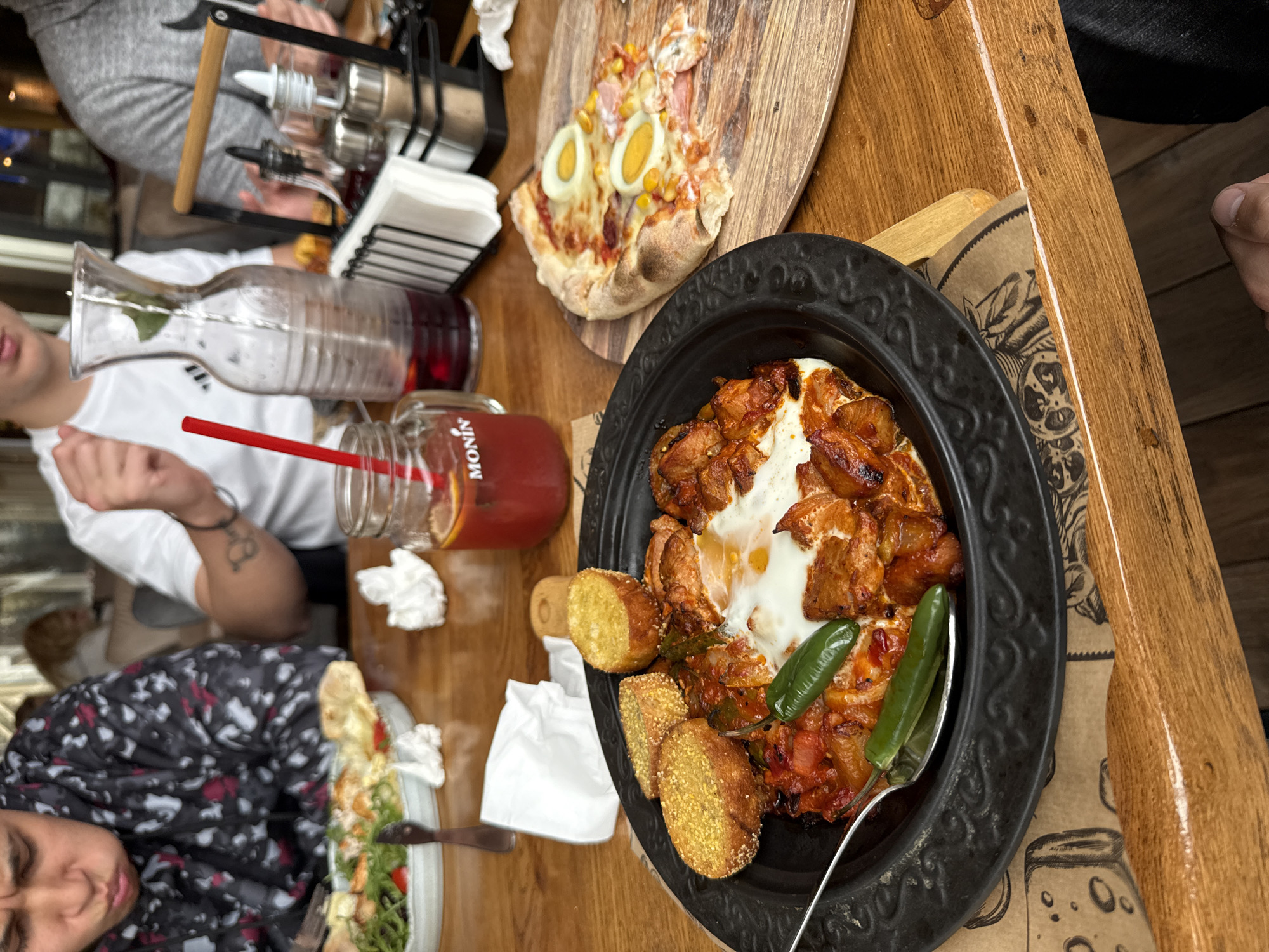 The image shows a wooden table set with various dishes, likely in a restaurant. A black, ornately designed plate contains a dish featuring cooked chicken pieces, a white sauce (possibly yogurt or labneh), fried bread slices, and green chilies. A silver spoon is placed in the dish. Next to it is a mason jar filled with a reddish-brown liquid, with the "Monin" brand visible. A pizza with boiled eggs and corn toppings rests on a wooden serving board. Further in the background, there are containers for spices and oil/vinegar in a black metal stand. Three people are partially visible around the table. The location is Unknown, Unknown.