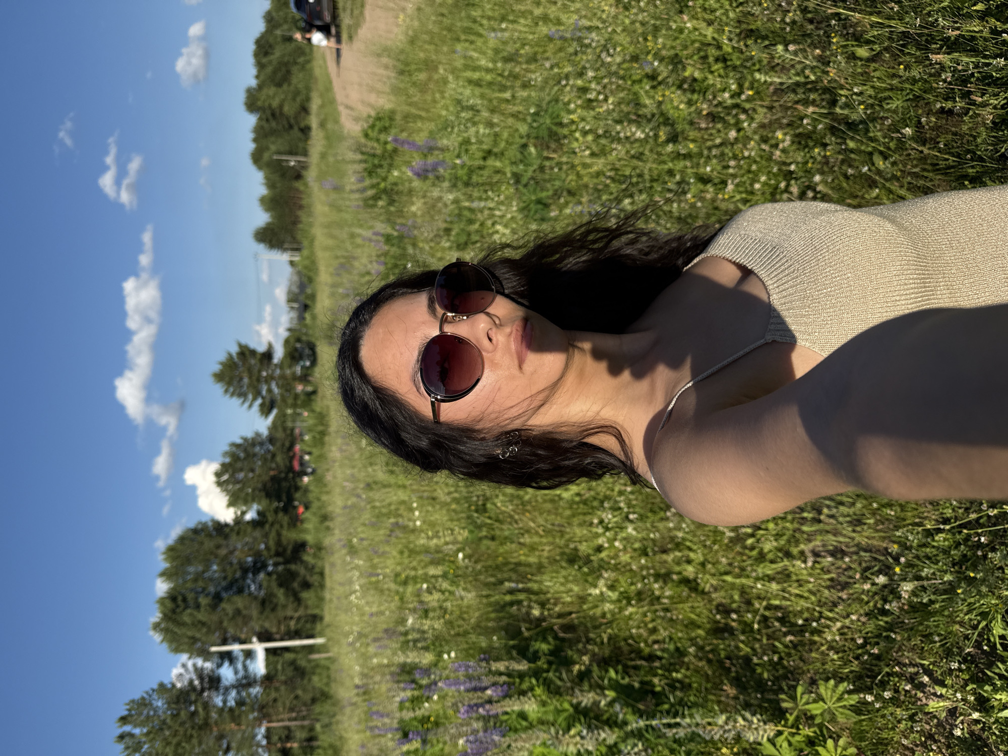 A woman with long, dark hair and sunglasses is lying down in a grassy field with purple flowers. She is wearing a beige, ribbed tank top. The sun is shining, casting shadows on her arm and face. In the background, there are trees and a blue sky with white clouds. There is also a dirt path and a few vehicles visible in the distance, suggesting a park or recreational area. The overall mood is relaxed and summery. The image is taken from a high angle, looking down at the woman.