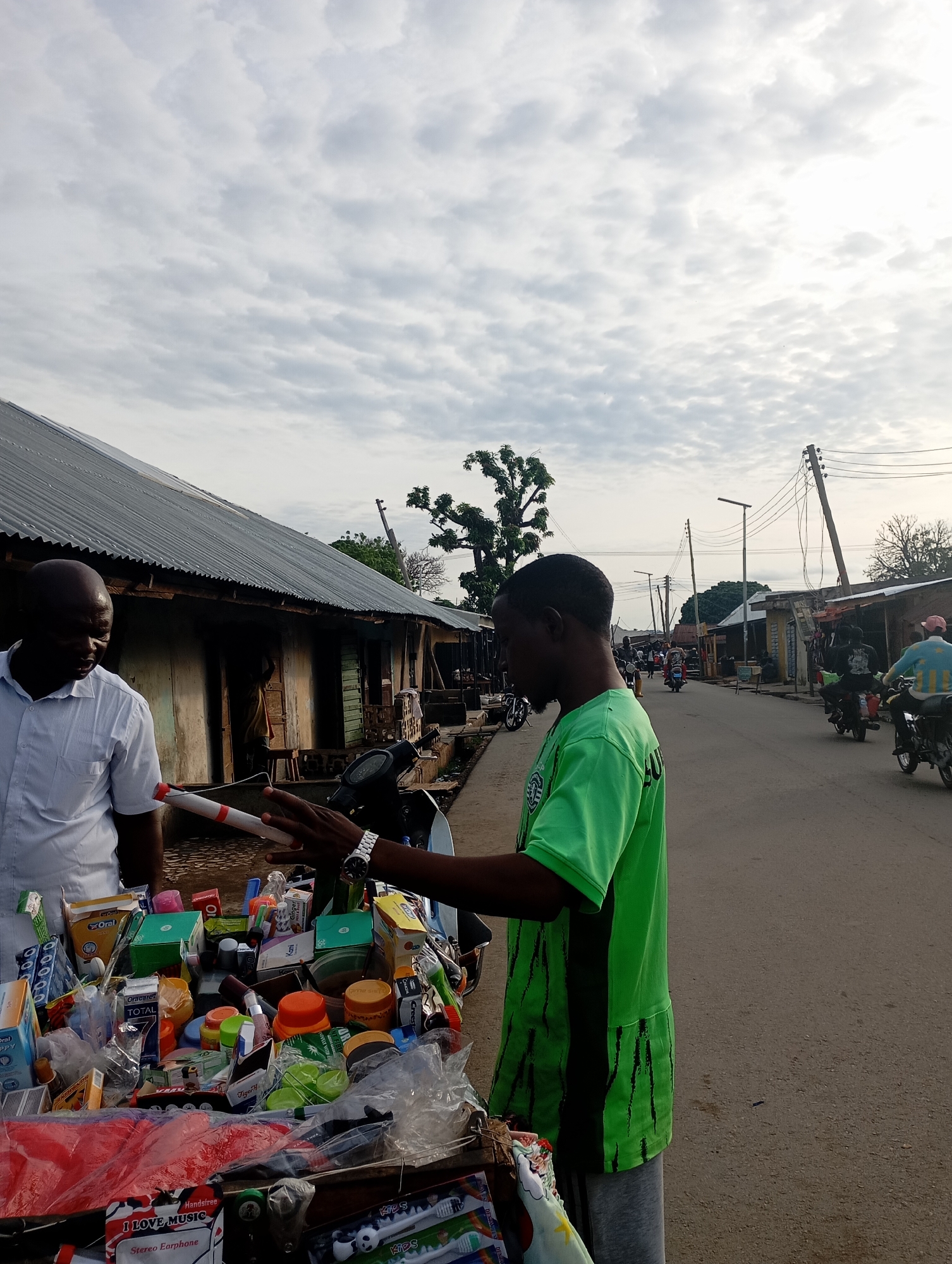 In Shendam, Nigeria, two men are interacting beside a cart laden with various goods. One man, wearing a light blue striped shirt, stands on the left, facing the cart. The second man, dressed in a bright green t-shirt with black vertical stripes and grey sweatpants, is on the right, actively gesturing towards the cart. The cart displays a colorful assortment of items, including boxed medications, toiletries, small containers, and bagged products. A motorcycle is parked behind the cart, partially visible. The scene unfolds on a paved street lined with buildings constructed with corrugated metal roofs and stucco walls, suggesting a commercial or residential area. The sky is overcast with a pattern of textured clouds. In the background, other individuals and motorcycles are present, indicating ongoing street activity.
