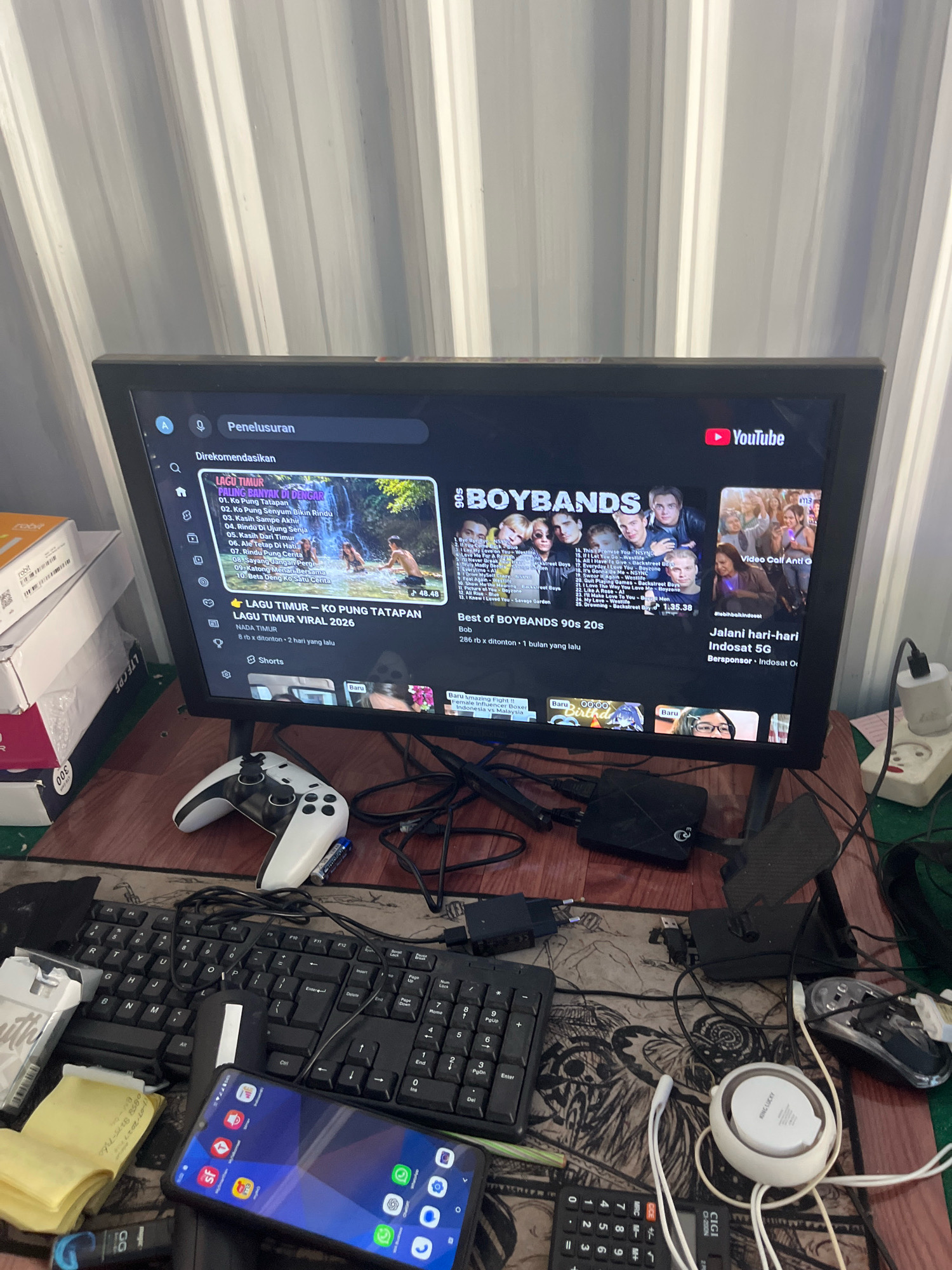 The image depicts a desk with a computer monitor displaying YouTube. The monitor shows a search page with recommendations for "LAGU TIMUR" and "BOYABNDS". A white video game controller is placed in front of the monitor. To the left of the monitor, there are stacks of boxes. On the desk surface, a black keyboard and a mouse are visible, along with a smartphone displaying app icons. Various cables and electronic devices are scattered around the desk. In the background, a white ribbed wall is present. The scene appears to be an indoor setting, likely a room in Sengkang, Indonesia, given the contextual information. There are no people visible in the image.