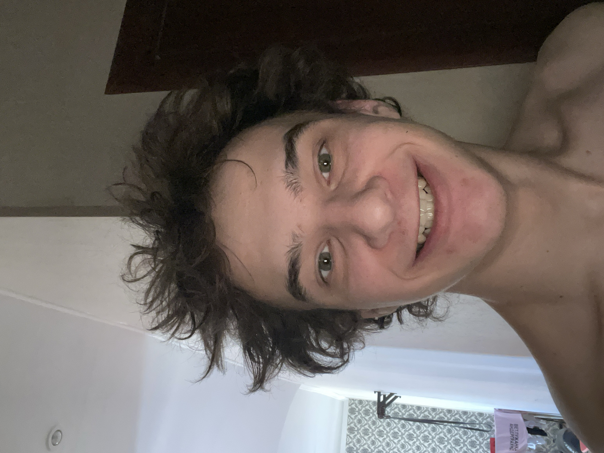 The image features a young man, likely in his late teens or early twenties, captured in a selfie. His head is tilted slightly to his right, and he is looking directly at the camera with a wide, cheerful smile that reveals his teeth. He has green eyes and dark, wavy, somewhat disheveled hair. He appears to be shirtless, with his bare shoulder and upper chest visible.
The setting is indoors, characteristic of a personal room in Krasnoyarsk, Russia. In the upper part of the background, a dark, possibly wooden surface or bed frame is visible above his head. Below him, a light-colored ceiling or wall occupies the upper left section of the frame. Towards the lower right, a wall covered in a repeating white and grey geometric patterned wallpaper can be seen. A small, round, recessed light fixture is also visible on the light-colored surface.
In the bottom right corner, a pink or purple rectangular object, possibly a bag or box, rests against the patterned wall. Cyrillic text is visible on this object, which reads "ВЕТУХАНА АНДЕГРАУНД" (VETUKHANA UNDERGROUND). The lighting is soft and even, suggesting an indoor light source. The man's expression conveys happiness and playfulness.
