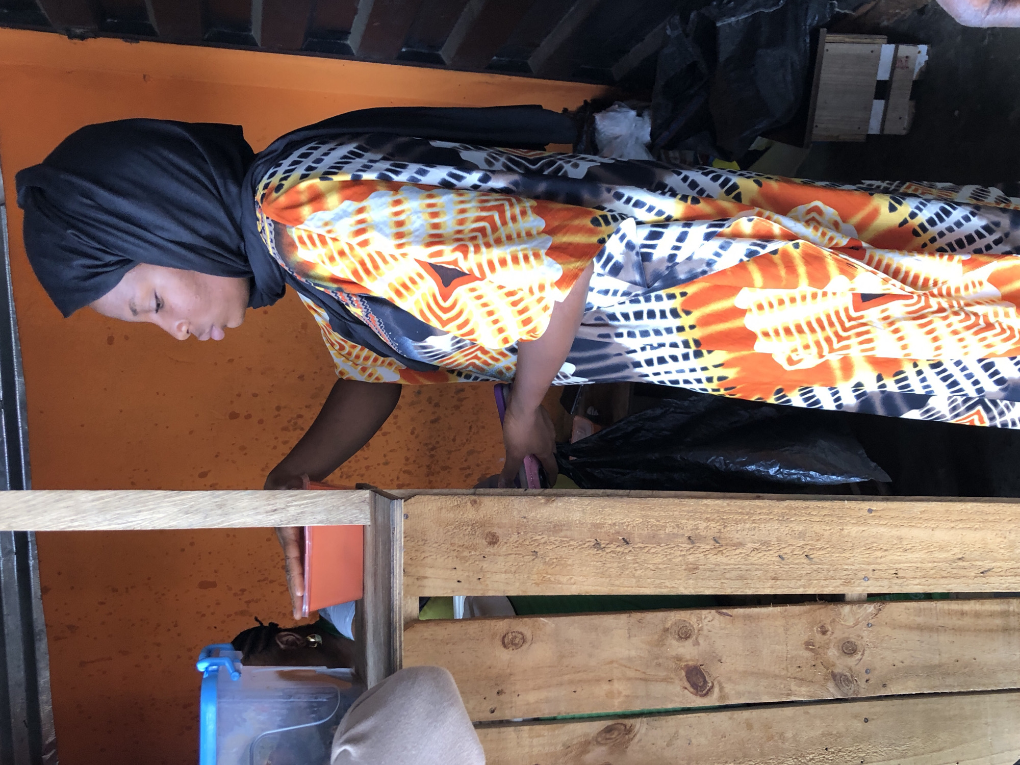 A young woman wearing a black hijab and a colorful, patterned dress stands in profile. She appears to be interacting with an orange plastic container. To her left, a clear plastic container with a blue lid is visible. The background is an orange wall. The scene is set outdoors, possibly at a market stall, given the wooden structure in the foreground and the presence of various containers and black plastic bags. The image suggests a casual, everyday moment in a bustling urban environment.