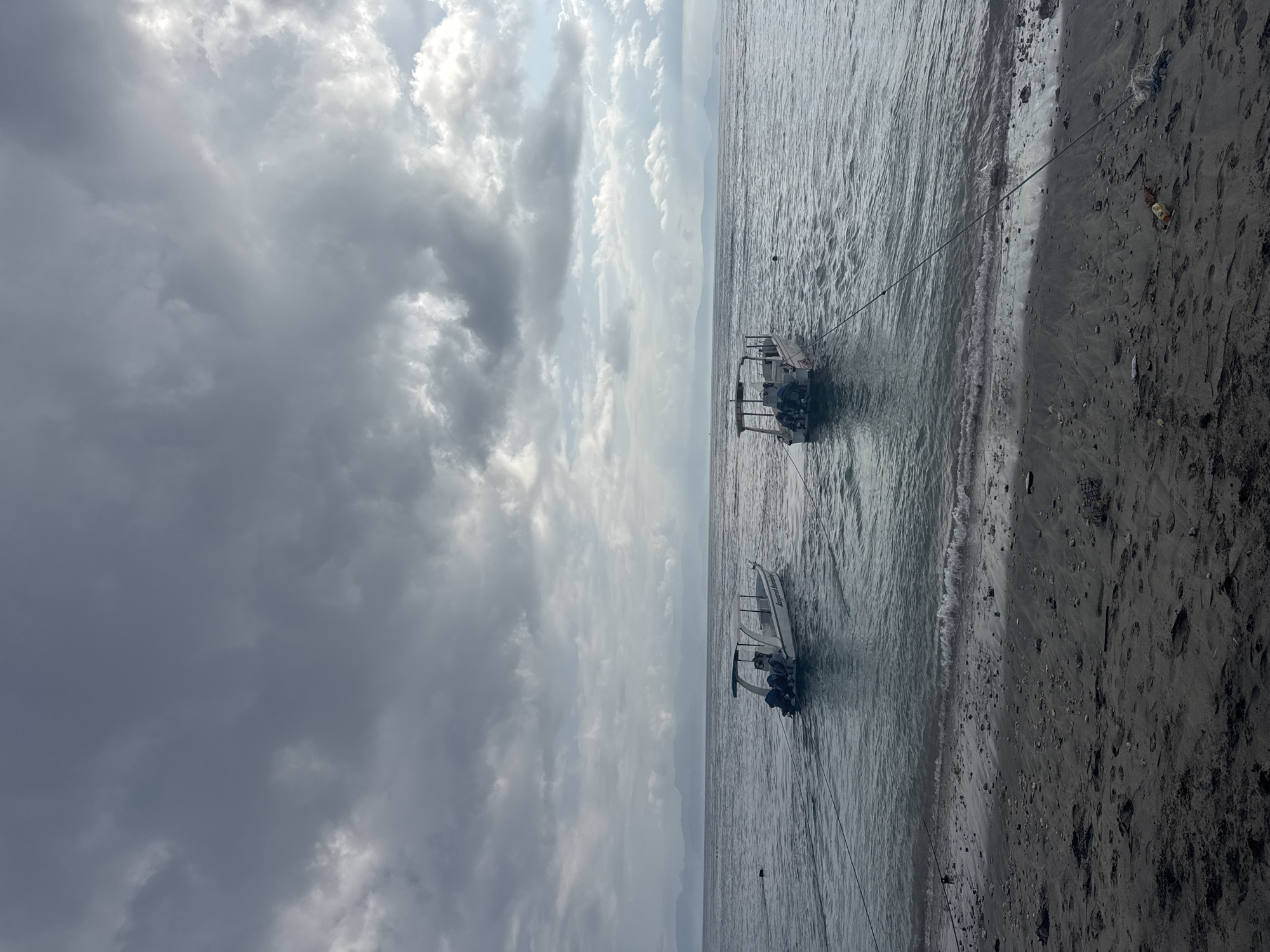 The image depicts a serene beach scene in Nusa Penida, Indonesia, under a dramatic, overcast sky. Two white speedboats, each equipped with twin outboard motors and a canopy, are anchored in the calm, grey water. They are positioned in the mid-ground, with one slightly closer to the shore than the other. The boats are tethered by ropes that extend towards the foreground, implying they are moored from the beach. The shore itself is composed of dark sand, strewn with pebbles and some debris, including a plastic bottle and a small piece of netting. Gentle waves lap at the shoreline. In the distance, a hazy mountain range forms the horizon, adding a sense of depth to the landscape. The overall atmosphere is one of quiet anticipation, with the cloudy sky suggesting a possible change in weather.