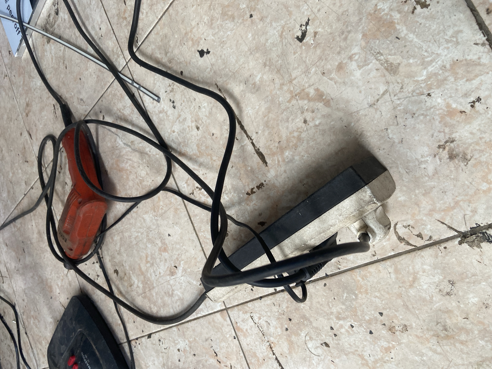 An overhead shot shows an assortment of electronic devices and cords strewn across a tiled floor. In the upper left corner, a black electronic device with red buttons is partially visible. To its right, a bright orange, corded electric drill lies on its side, its cord tangled. A power strip, predominantly white with a black strip along its top, is in the foreground, with several black cords plugged into it and draped around it. A thin, silver antenna-like object extends from the upper right of the frame. The floor tiles are a light beige color with a pinkish-brown marbled pattern, and the surface is dusty and appears to have some dark smudges and debris scattered across it. The scene is set in a room in Plosokandang, Indonesia.