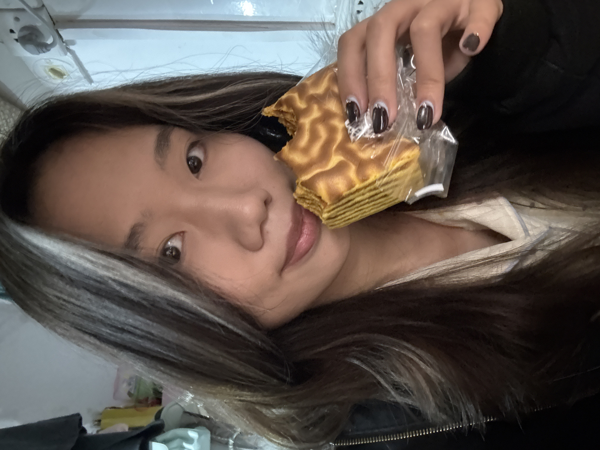 An individual, identified as a young Asian female, is depicted in a close-up, chest-up indoor shot. She has long, dark brown hair with lighter streaks and is looking directly at the camera with a slight smile. She wears a dark jacket over a lighter garment, possibly white or cream-colored, with light blue piping. Her fingernails are manicured with dark polish featuring white crescent designs at the base. She holds a partially consumed, layered golden-brown food item, resembling a cake or pastry, wrapped in clear plastic, in her right hand. A bite has been taken from the upper right portion of the food. The background to her left reveals partially visible hanging dark clothing and other indistinct objects, including a yellow item and a green container. To her right, a white wall features a light-colored electrical outlet or switch plate with a plug, and a vertical white element, likely part of a door frame or furniture. The specific location within Nan'an District, China, is not visually discernible from the image; however, the scene depicts an interior setting, likely a private residence or room.