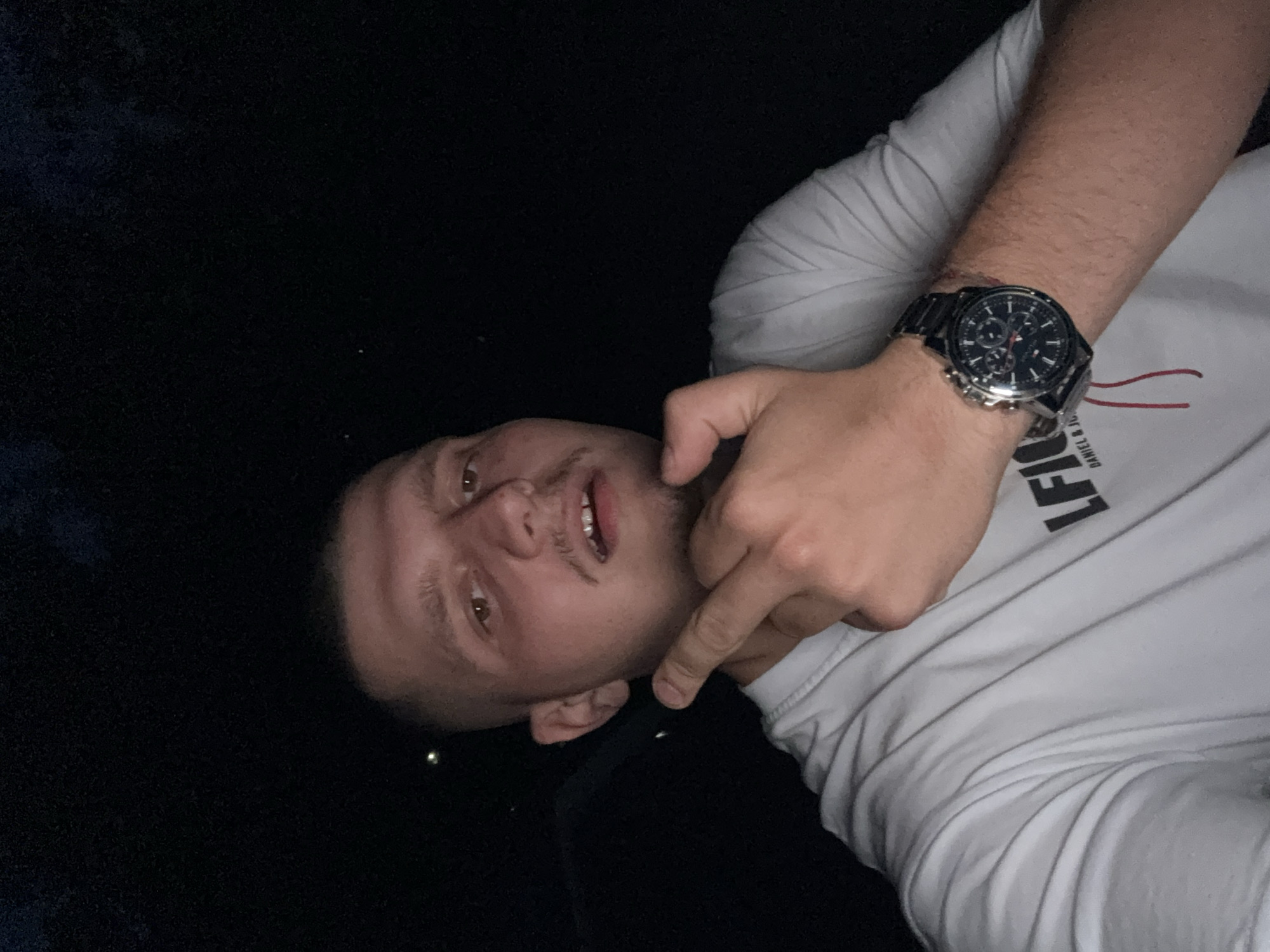 A male individual, appearing to be in his twenties or early thirties, is depicted in a close-up portrait, looking directly at the camera. He has short, light-colored hair, a light mustache, and stubble. He wears a white, short-sleeved t-shirt with a dark logo (partially visible "LFI..." and "DANIEL & JO...") on the chest. His left hand is raised to chin level, with the index finger bent and the thumb raised, positioned near his lower jaw. A dark-colored chronograph watch with a metal band is worn on his left wrist, and a thin red string is tied below it. The background is completely dark, indicating a nighttime setting or a severely unlit interior, with a few faint, blurry light points visible in the upper corners. The subject is illuminated by a direct frontal light source, likely a camera flash. The scene is situated in the city of Blagoevgrad, Bulgaria; however, the obscured background provides no specific topographical or architectural details to identify the precise location within the city.