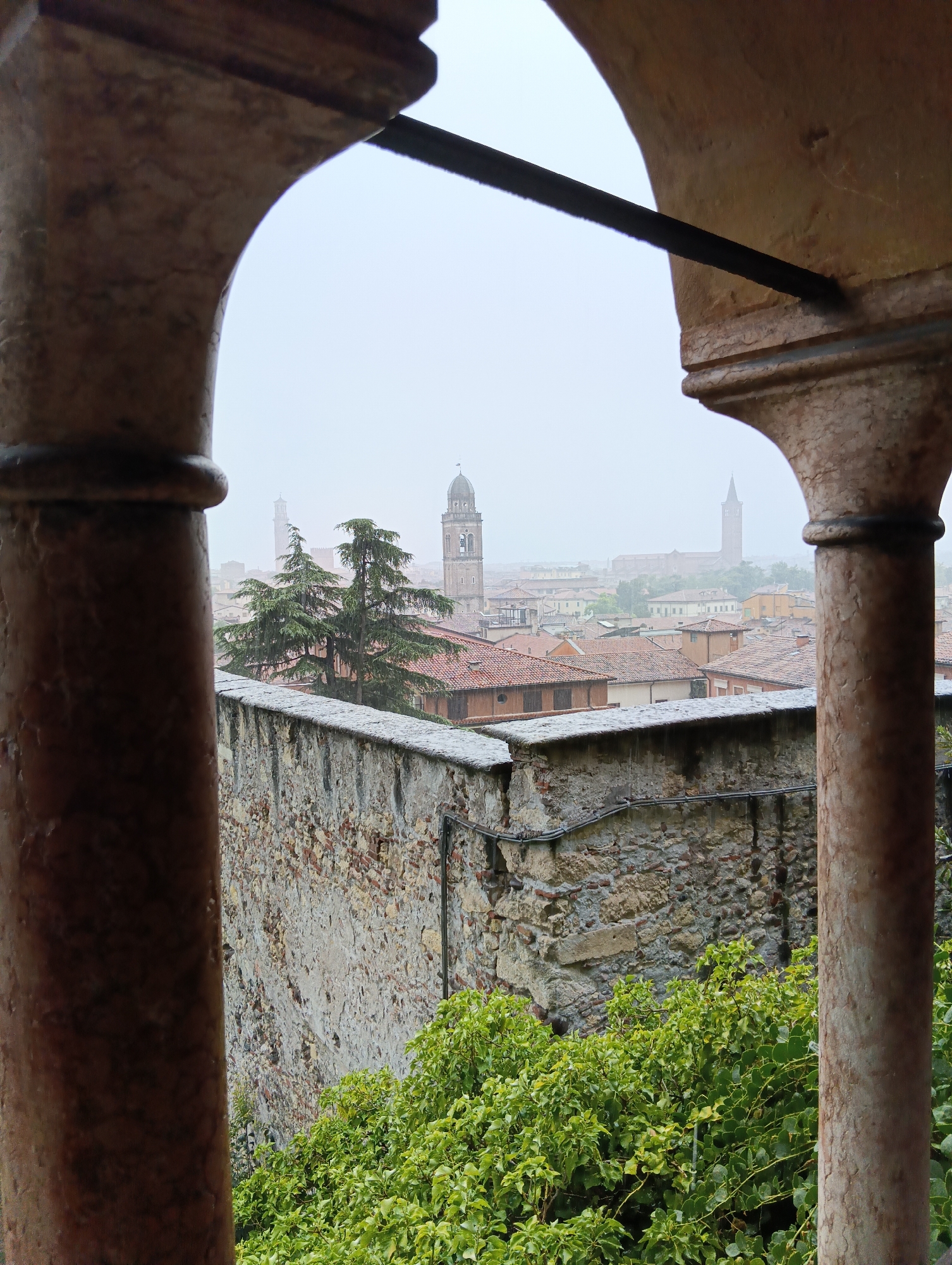 The image shows a view of Verona, Italy, through an arched stone opening. The foreground features weathered stone pillars and a stone wall topped with greenery. Beyond the wall, the city skyline is visible, characterized by red-tiled roofs and several church towers. The sky is overcast, suggesting a rainy or foggy day, with rain visible in the air. In the mid-ground, two evergreen trees stand prominently. The architecture and church towers suggest this is a historical part of the city. There are no people visible in the image.