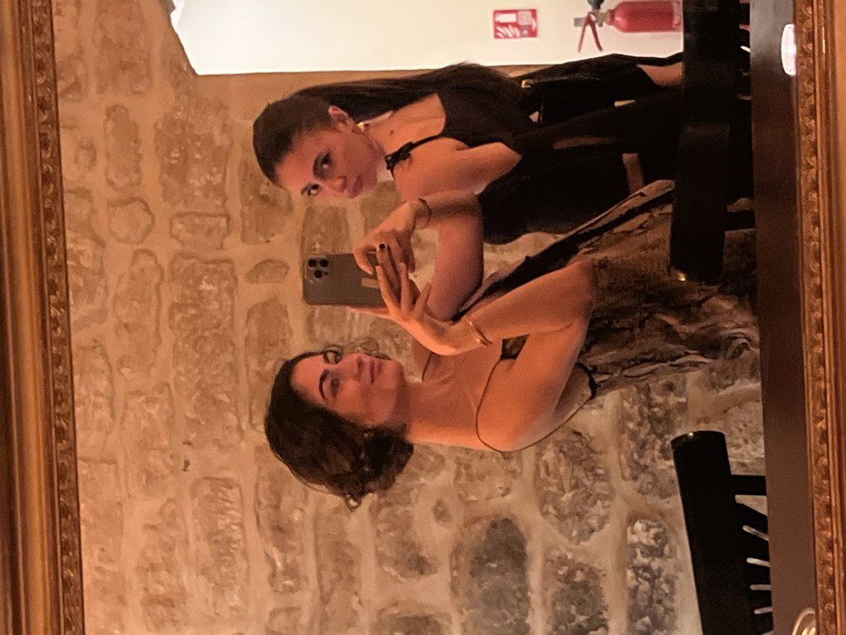 A mirror reflects two women against a stone wall. One woman, with dark hair pulled back, wears a black dress and looks towards the camera. The other, with shorter dark hair, holds a phone and appears to be taking a selfie. A fire extinguisher and exit sign are visible in the upper part of the reflection. The mirror has an ornate gold frame, partially visible on the edges. A black chair or object is also reflected, adding depth to the image.