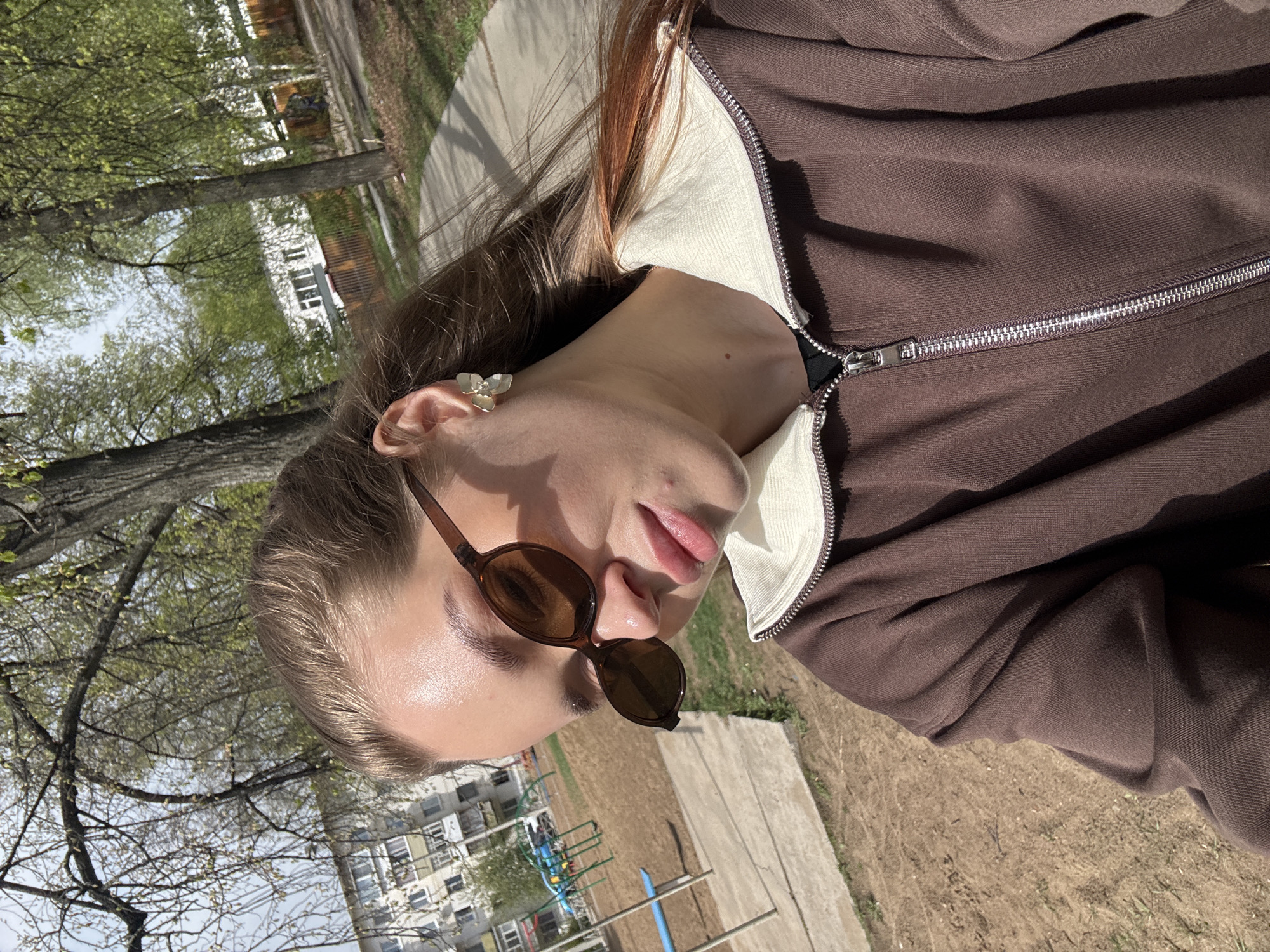 A person with light skin and brown hair is outdoors, wearing sunglasses and a brown zip-up jacket with a light-colored collar. The surroundings include trees and a playground, suggesting a park or recreational area in the city of Chaikovsky, Russia. A building is visible in the background.