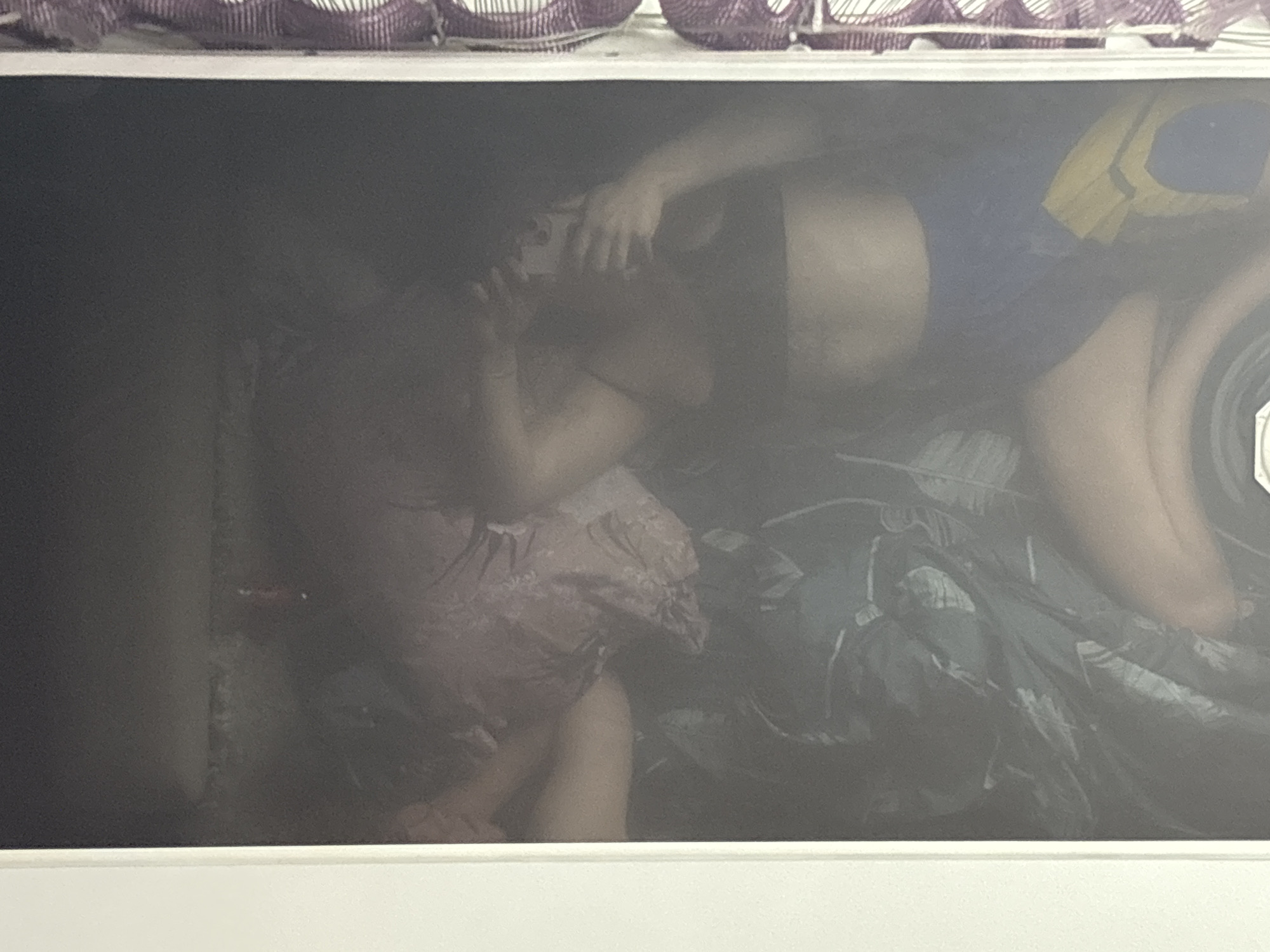 The image appears to be a photograph of a framed print or artwork, possibly taken in a dimly lit environment. The subject matter within the frame depicts two individuals lying on what seems to be a bed or surface covered with bedding.
One individual is wearing what appears to be a pink or patterned dress and is positioned on the left side of the frame. The second individual is wearing dark shorts and a blue and yellow shirt and is positioned to the right. Their arms are raised, and they may be interacting with a phone or other device. The background is obscured, but the environment appears to be a bedroom or similar indoor space.
The location (Perm, Russia) is not visually apparent in the image.