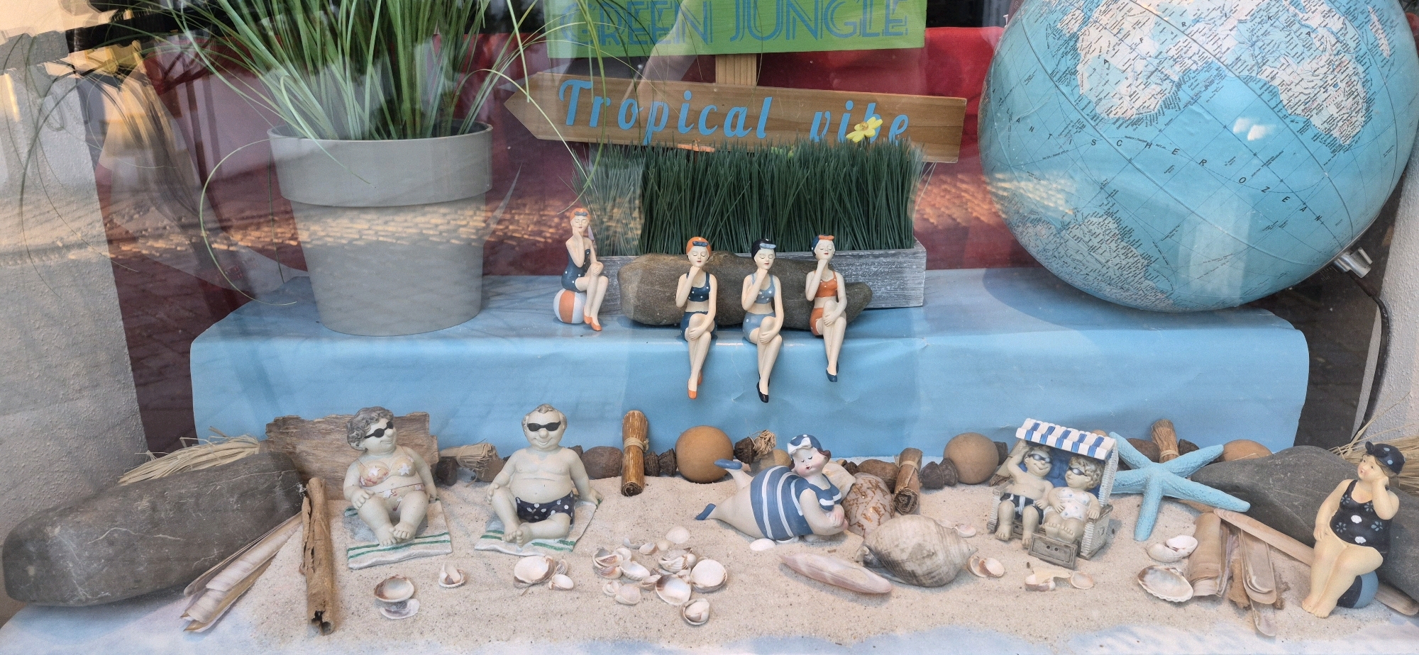 The image depicts a shop window display, likely for a gift shop or novelty store, in Ingolstadt, Germany. The display has a beach theme. In the background, there is a large blue globe and a potted plant with long green leaves. A wooden sign reads "GREEN JUNGLE" and "Tropical vibe" with a small yellow flower.
The main focus of the display is on the sand-covered surface in the foreground, which is populated with several ceramic figurines. On a raised blue platform in the middle ground, three female figurines in vintage swimwear are seated on a rock. In the foreground, on the sand, there are two larger figurines of a woman and a man, both in swimwear and sunglasses, lying on towels. Another figurine of a person in a striped swimsuit is partially submerged in the sand. To the right, two children are seated under a beach umbrella, and a woman in a polka dot swimsuit is sitting on a ball. The sand is scattered with seashells and driftwood. The display appears to be set up to evoke a summer vacation or beach atmosphere.