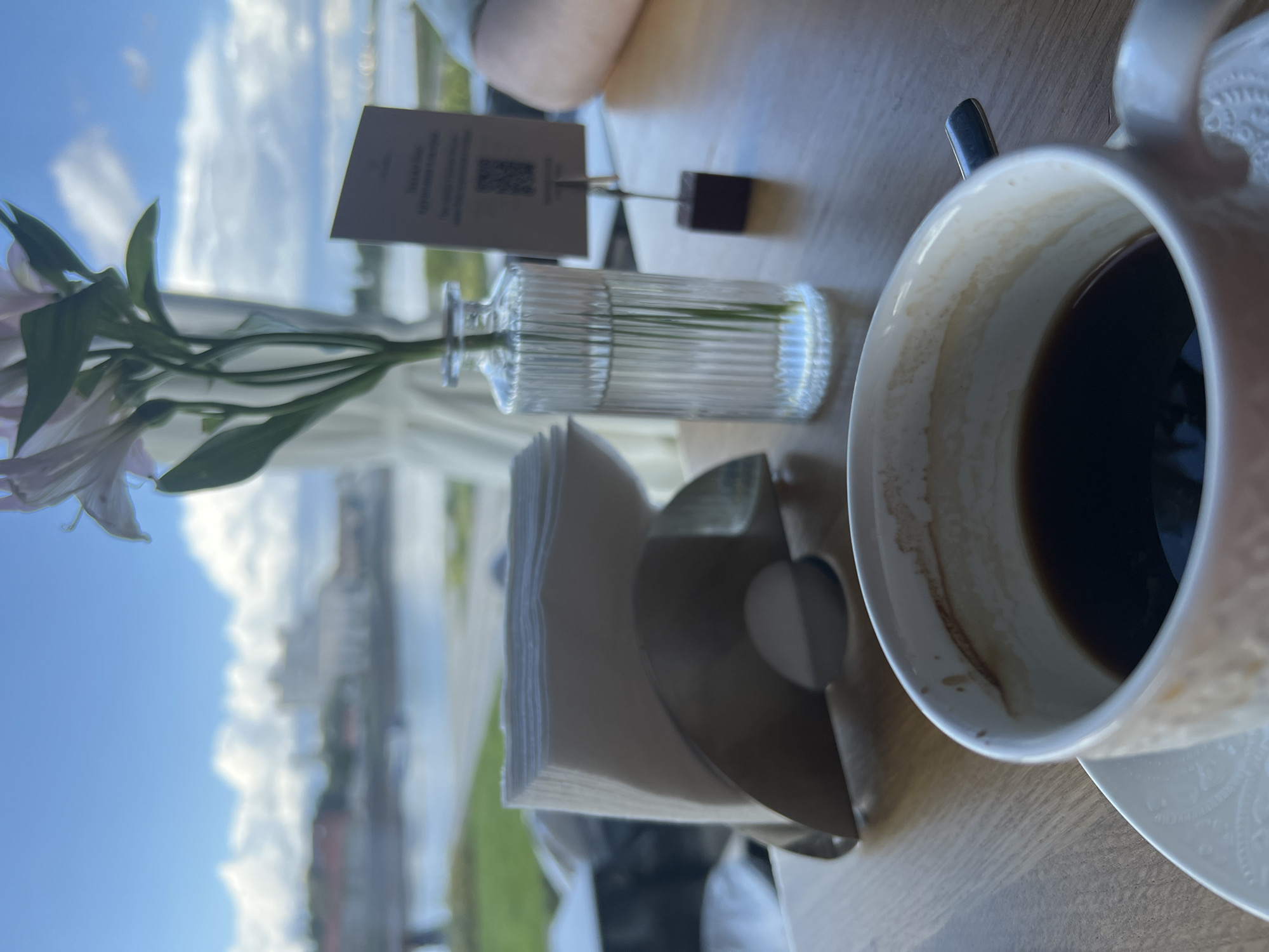 A cup of coffee rests on a saucer on a wooden table, with a stack of napkins to the left and a vase with flowers behind it. In the background, a body of water is visible, with buildings and a cloudy sky beyond. The scene appears to be set in a cafe or restaurant in Veliky Novgorod, Russia, given the architectural style of the background buildings. There are no visible people or actions.