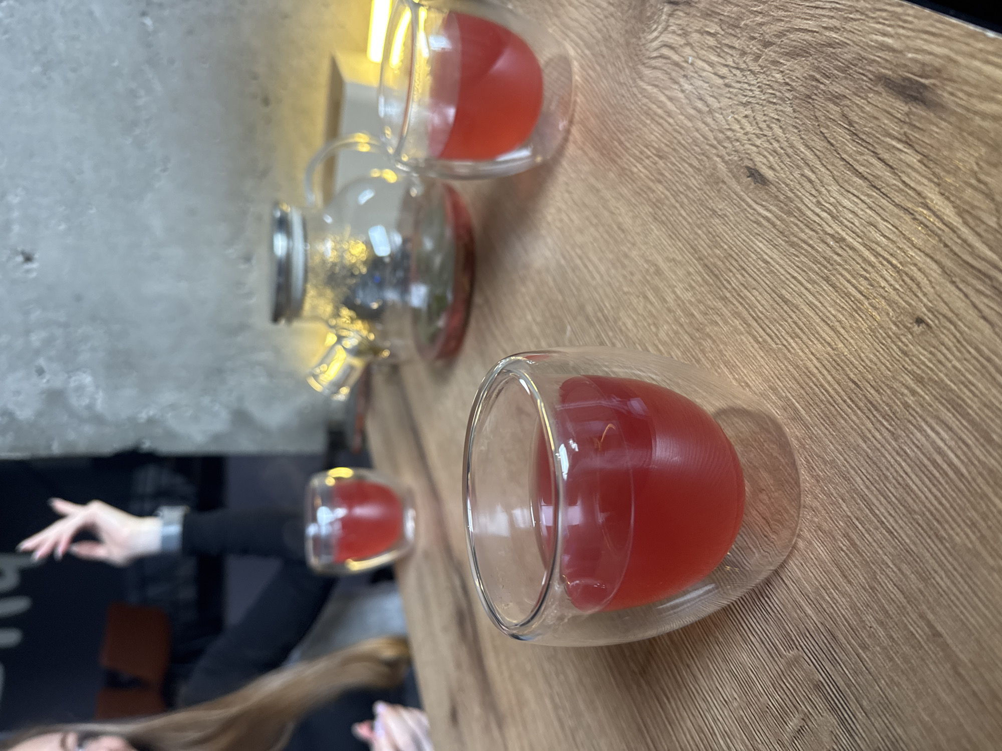 The image captures a scene featuring three glasses of red liquid and a glass teapot, likely filled with tea, set on a wooden table. The location appears to be a café or restaurant, possibly in Unknown, Unknown, based on the background with the partial view of a person in a black outfit and blonde hair. The glasses are arranged along the table's surface, suggesting a casual or social setting, and the presence of the teapot implies that the red liquid is being served or shared. The wooden table has a prominent grain pattern, and a partial view of a person is visible, indicating interaction within the environment.