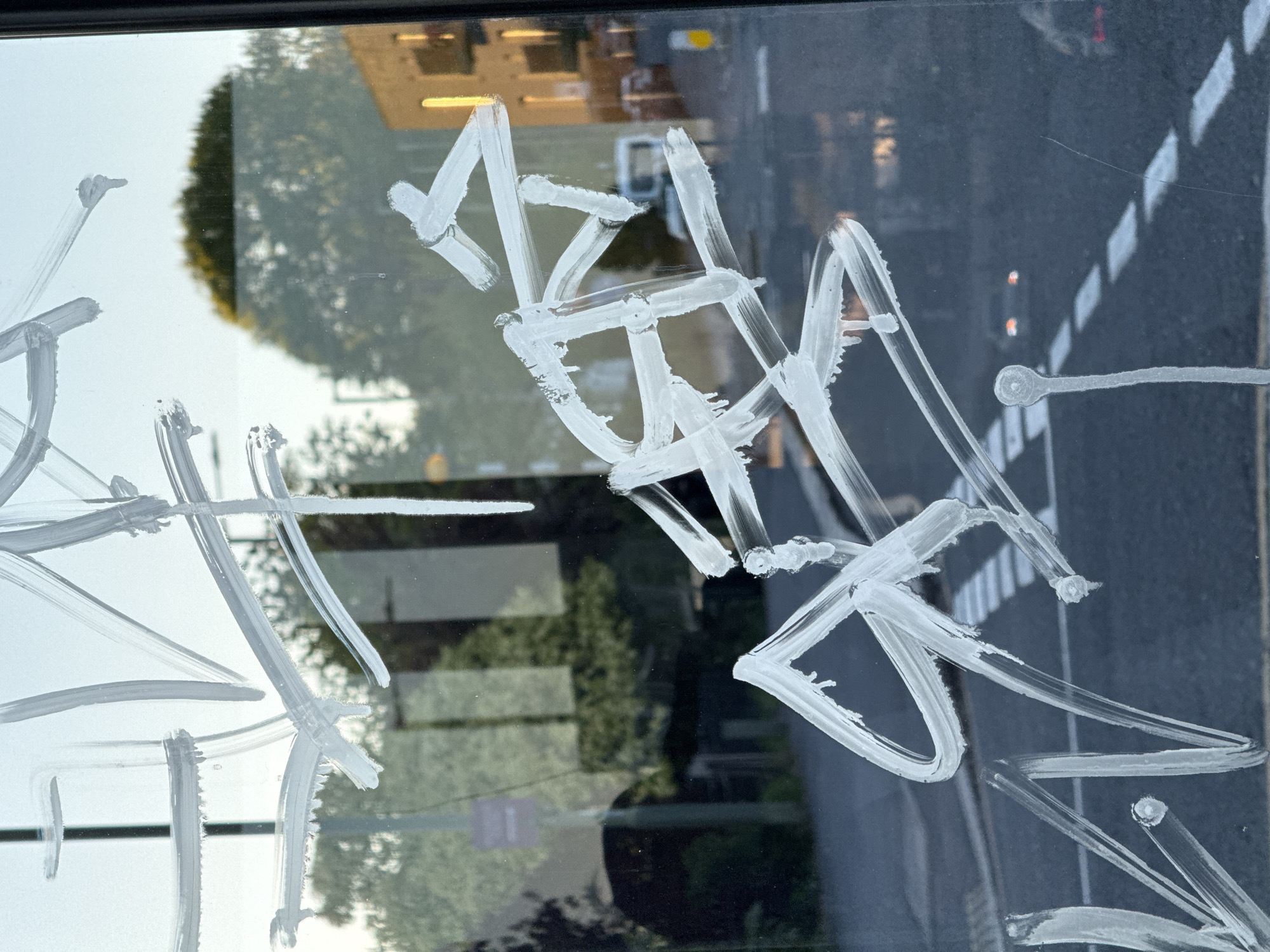 The image presents a close-up, slightly tilted view through a window that has been marked with thick, white, abstract writing, likely applied with paint or a similar substance. The markings are large, stylized, and appear to be a form of graffiti or tag, characterized by bold strokes, sharp angles, and overlapping lines. While specific words are difficult to decipher, individual elements resemble letterforms, with a prominent 'M' or 'A' shape visible in the upper central area, and various other intricate, unreadable symbols extending across the glass.
The window reflects an outdoor urban or suburban environment, consistent with a London, United Kingdom setting. The reflections reveal a clear daytime scene, with bright light illuminating the top left quadrant, suggesting a clear sky. A large, leafy green tree is prominently reflected on the left. On the right side, the reflections are darker and show a road with distinct white markings, possibly indicating a zebra crossing or lane divisions, extending into the distance. Faint lights and blurred architectural structures are also visible in the background, suggesting buildings lining the street. No people or specific events are directly visible, but the reflections imply a typical street scene. The time of day appears to be daylight hours, with ample natural light.