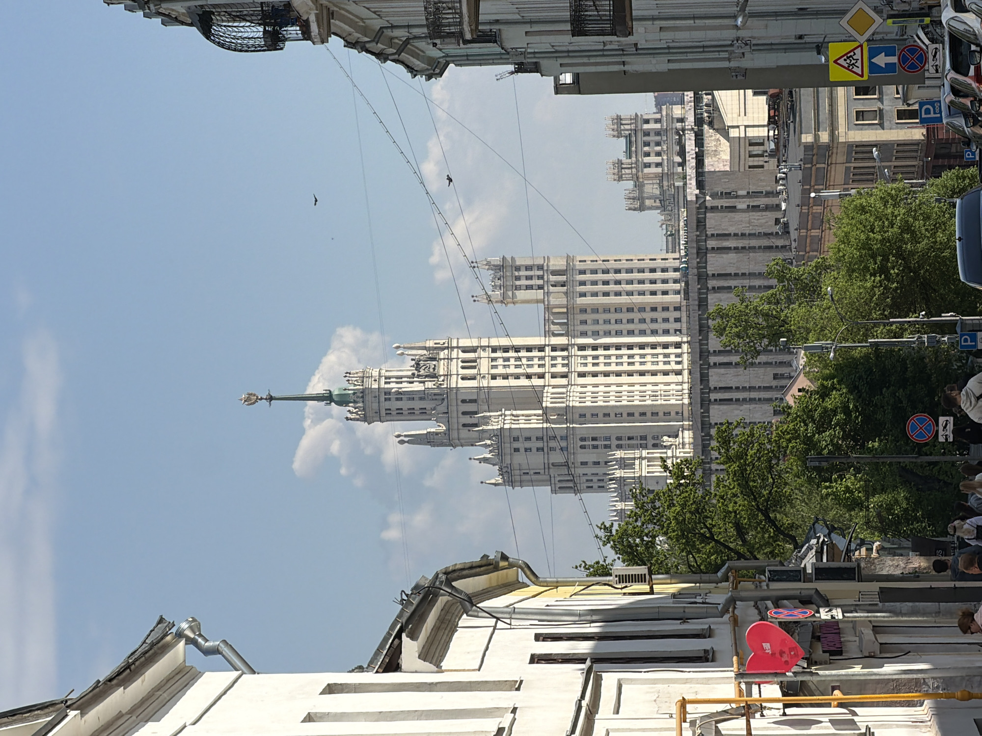 A vertical eye-level medium shot shows a view of the Kudrinskaya Square Building in Moscow, Russia. The Stalinist skyscraper is centered in the frame, framed by buildings on either side. The sky is blue with some clouds. Overhead wires run across the sky in the foreground. A bird is visible in the sky above the skyscraper. Lower-rise buildings flank the skyscraper, interspersed with greenery. Sidewalks with pedestrians are visible on the right side of the image, along with road signs and parked cars. The buildings framing the skyscraper are white and cream-colored, with architectural details visible. A red, heart-shaped object is visible on a lower building in the foreground.