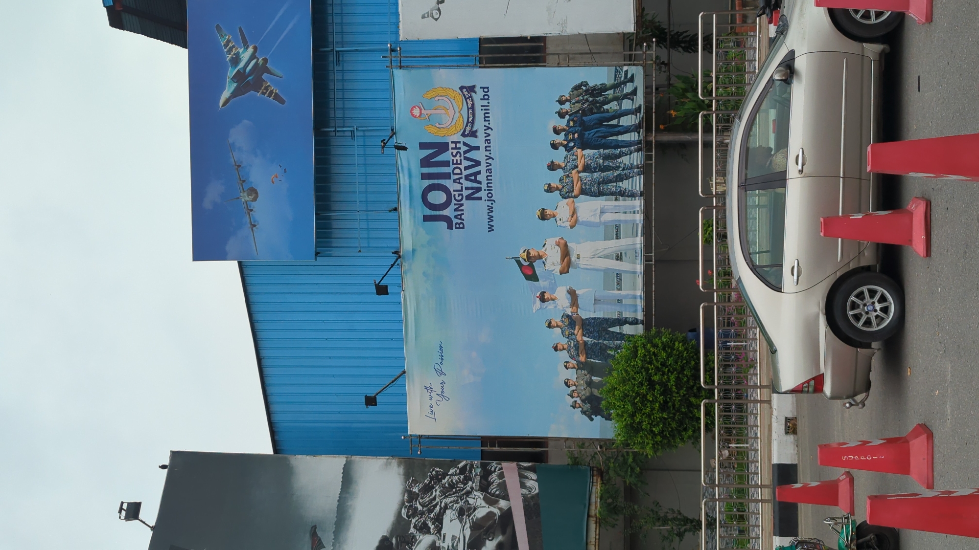 The image shows a street scene in Dhaka, Bangladesh. The focus is on a parked silver sedan surrounded by red traffic cones. Behind the car is a metal fence and some greenery. In the background are several billboards attached to a blue building. One billboard features fighter jets and a plane, another advertises the Bangladesh Navy with the website "www.joinnavy.navy.mil.bd," and a third shows an image of soldiers. The sky is overcast.