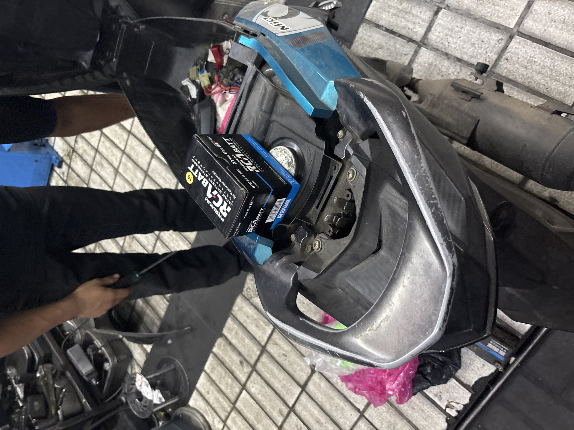The image shows a person in black pants working on a blue and black scooter in a workshop. The scooter's seat is open, revealing wires and a new battery box labeled "RCA BATT" and "GTZ-5S X GEL". A person's hand, holding a screwdriver, is visible on the left side of the image. The workshop appears to be tiled with light-colored squares and has various tools and equipment in the background. The scene suggests a battery replacement or repair is in progress. The location is likely a workshop in North Jakarta, Indonesia, given the context provided.