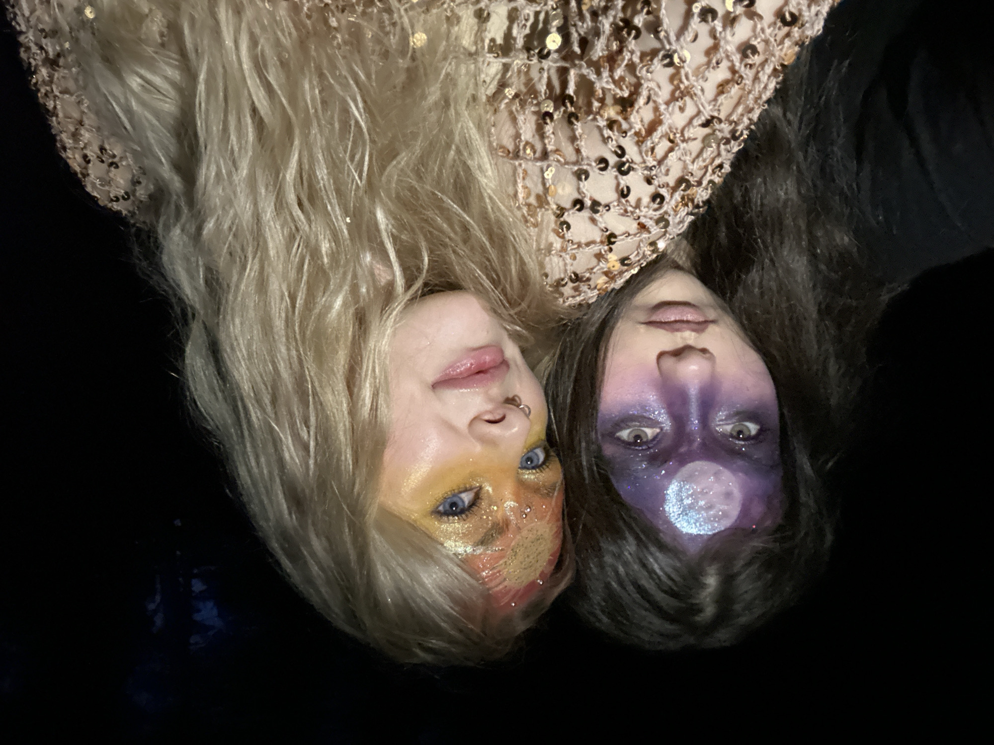 Two people with elaborate face makeup are lying head-to-head against a dark background. The person on the left has blonde hair and makeup featuring yellow, gold, and red glitter and a possible sun or flame motif on the face. The person on the right has dark hair and dark purple face paint with a large, round, sparkly design resembling a moon on their forehead. The top of the frame shows a textured, shiny garment or cloth, possibly adorned with sequins or small metallic discs. The location and city of Chaikovsky, Russia are not discernible.