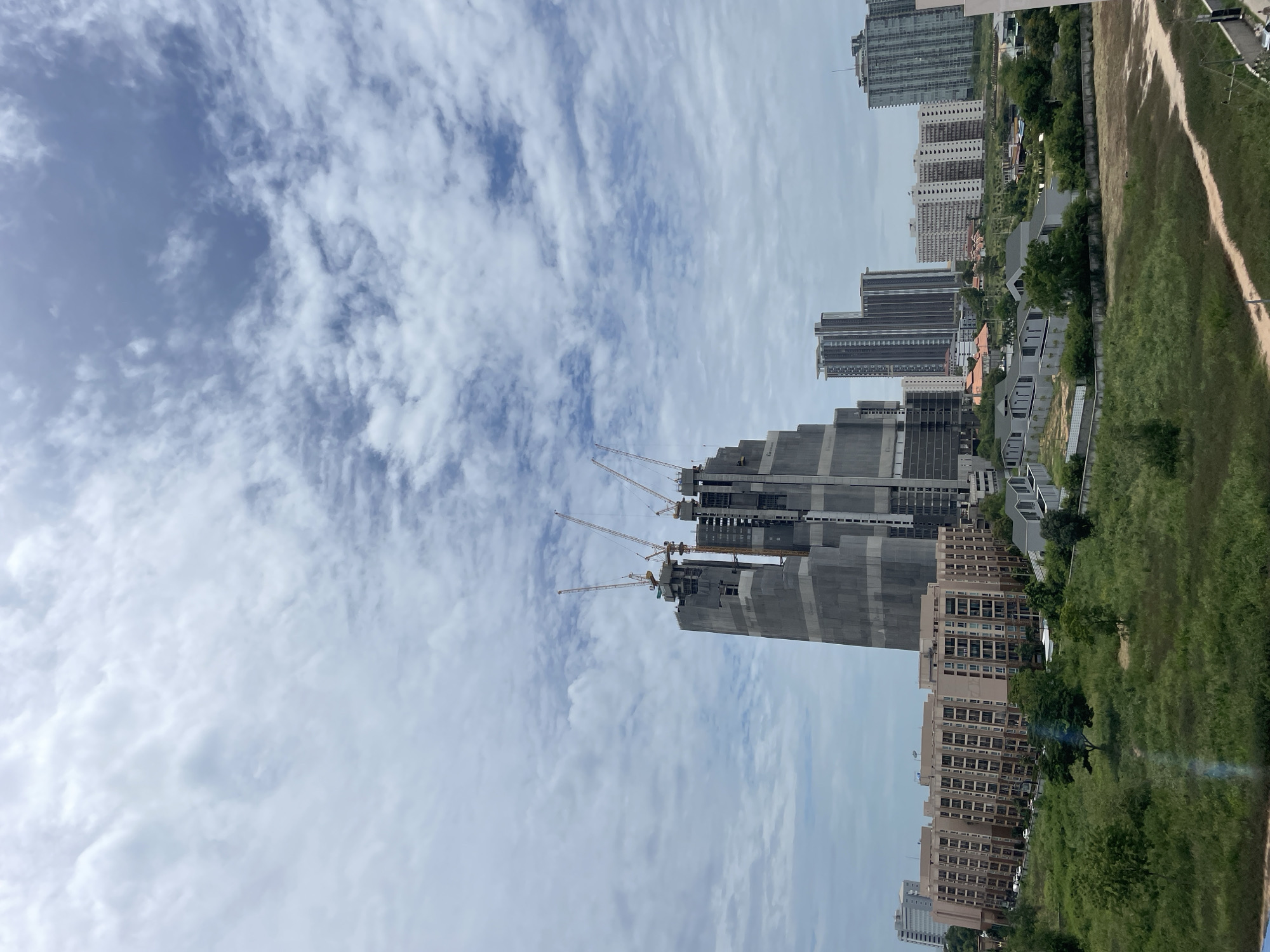 A vertical, medium-shot, outdoor, daytime image shows a city skyline of Pattaya City, Thailand, dominated by a massive, unfinished skyscraper that is covered in a grey tarp. Several yellow construction cranes are attached to the top of the skyscraper. In the foreground, there are several medium-rise apartment buildings, some of which are completed and others that appear to be under construction. The buildings are light brown and beige with many windows. In front of the apartment buildings is a green grassy area with scattered trees and a dirt path. The sky above is filled with a mix of white and grey clouds. The scene appears to be a developing urban area, with a blend of residential buildings and large construction projects.