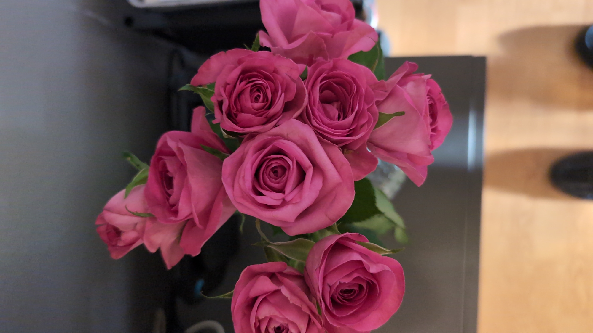 A bouquet of pink roses is displayed on a dark gray surface, possibly a table or shelf. The roses are in various stages of bloom, with some tightly closed buds and others more open, revealing their intricate petal structures. The lighting casts shadows on the surface below, and a wooden floor is visible in the lower portion of the image. Two blurry dark shapes, likely shoes, are present on the wooden floor, indicating a person's presence in the vicinity. The background is out of focus, suggesting a shallow depth of field or the focus being solely on the roses. There is no identifiable context that would place this scene in Worms, Germany.