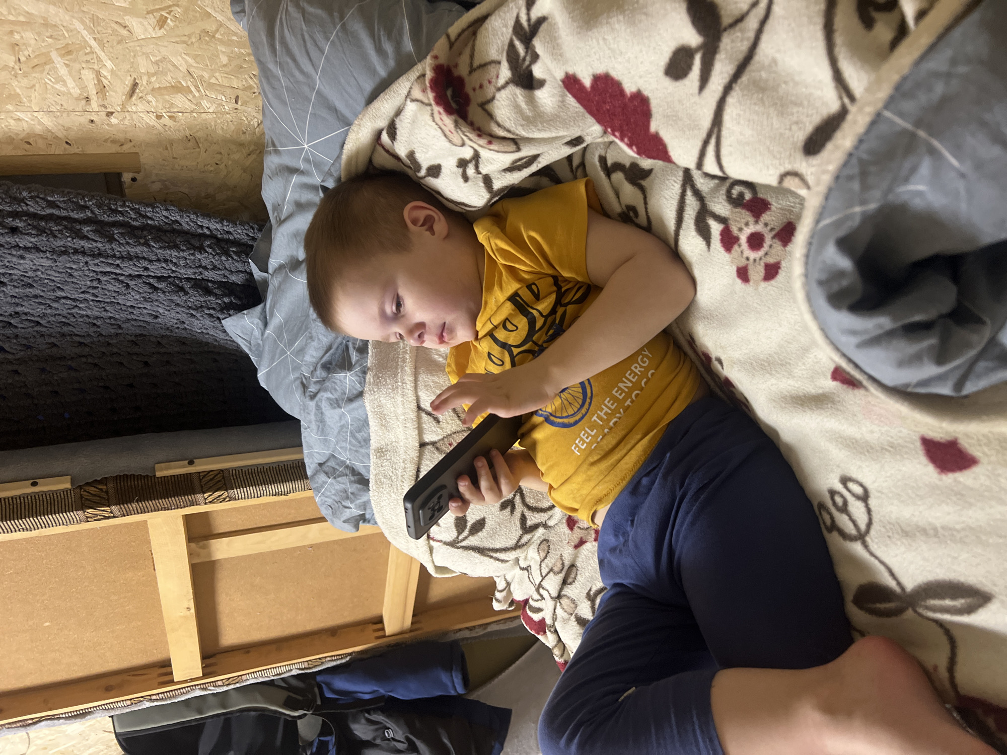 A young child, likely a boy, is lying on their left side on a bed indoors. The child has short brown hair and is wearing a yellow short-sleeved t-shirt with partial text "FEEL THE ENERGY" and "READY TO GO" visible, along with a bicycle-themed graphic. They are also wearing dark blue pants. The child is holding a black smartphone with both hands, appearing focused on its screen. A white and cream blanket with red and brown floral patterns partially covers the child, resting over a grey sheet with a light geometric pattern. The back of the phone case has a small white emblem with the number "33." The bed is situated against a wall covered with oriented strand board (OSB) paneling, and wooden bed frame elements are visible beneath the bedding. The child's expression is neutral and concentrated. The image was taken in Chaikovsky, Russia.