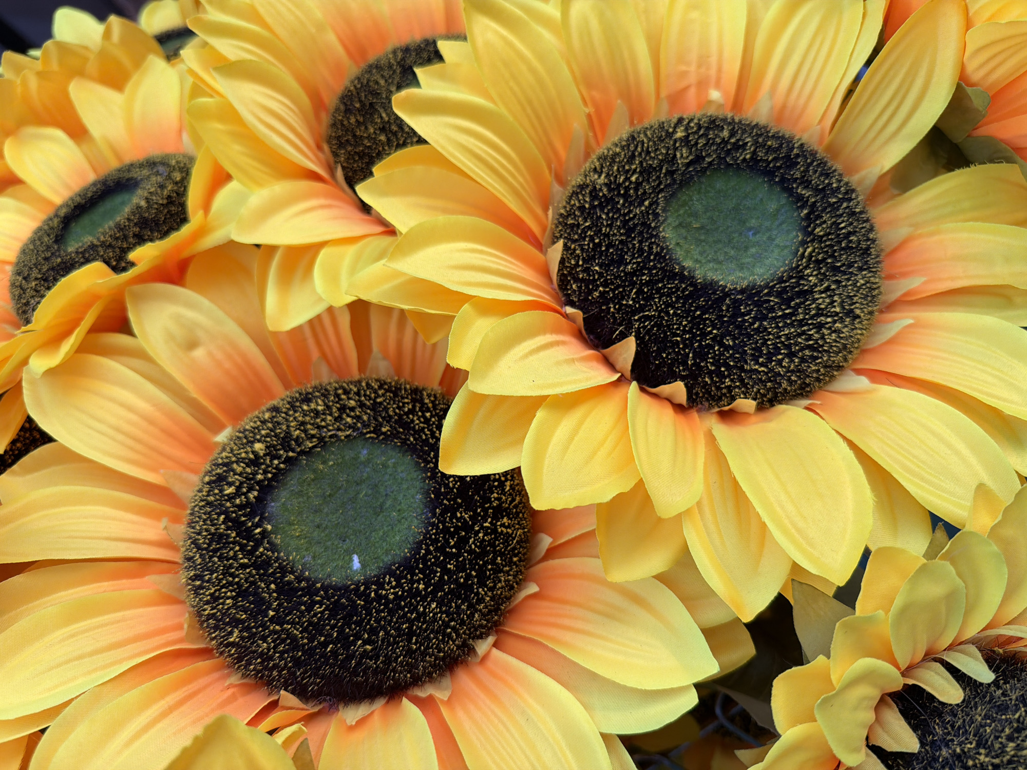 This is a close-up, overhead shot of a bouquet of artificial sunflowers. The flowers are vibrant yellow with orange-tinged petals closest to the dark, fuzzy centers. The centers appear to be a deep brown with a green, fuzzy core. The petals are layered and soft-looking, suggesting they are made of fabric. The bouquet is densely packed, with multiple flower heads overlapping each other. No people or specific locations within Alanya, Turkey, are visible in the image; it is a still life focused solely on the sunflowers.