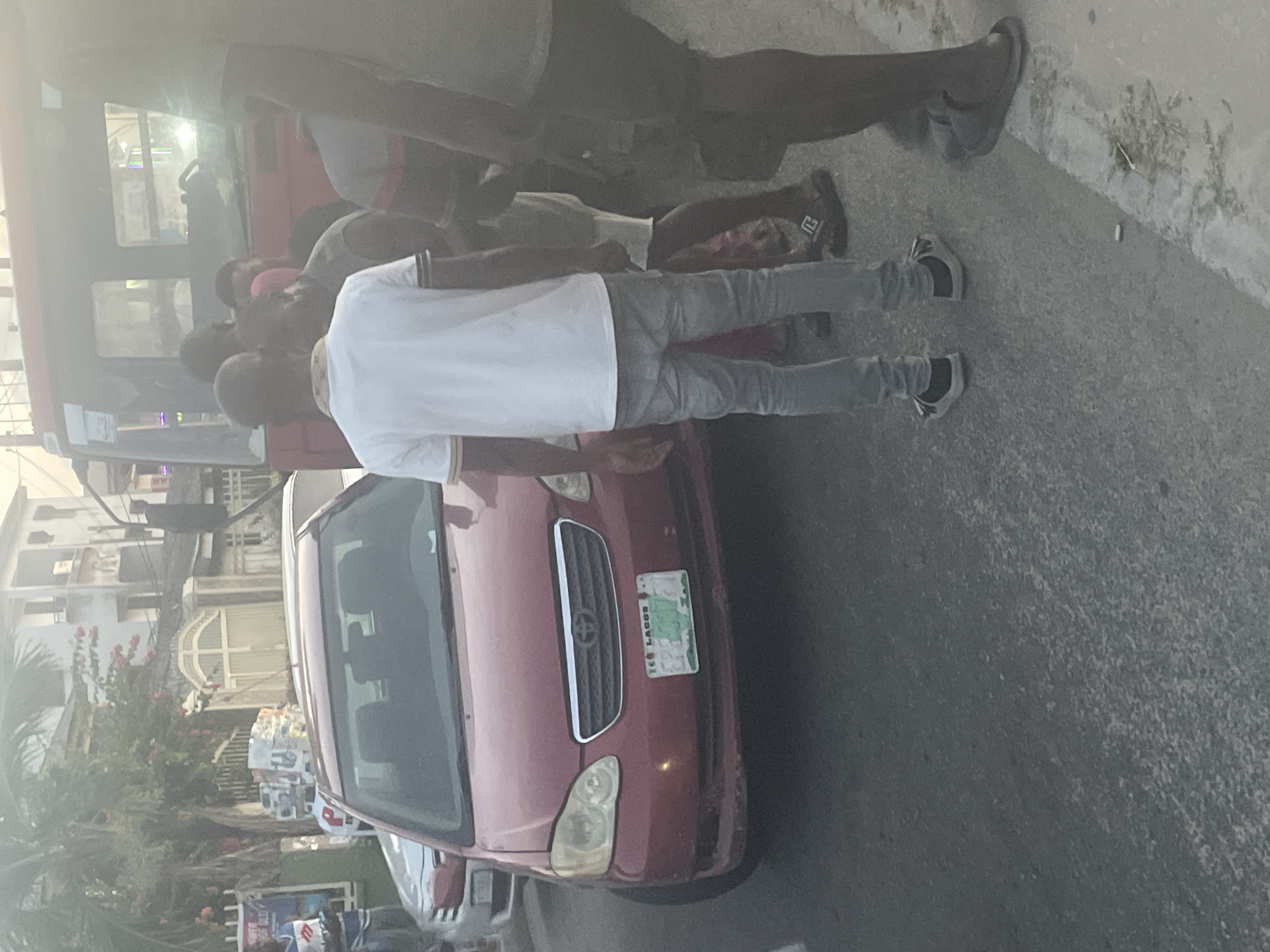The image depicts a street scene in Ado, Nigeria. A red Toyota Corolla is parked on the street, with a license plate reading "Lagos". Several people are gathered around the car and a nearby red bus. One man stands beside the car, wearing a white shirt and grey pants. Other pedestrians are visible, including one wearing flip-flops. In the background, there are buildings and what appears to be a vendor selling goods. The overall environment appears to be a typical urban street scene.