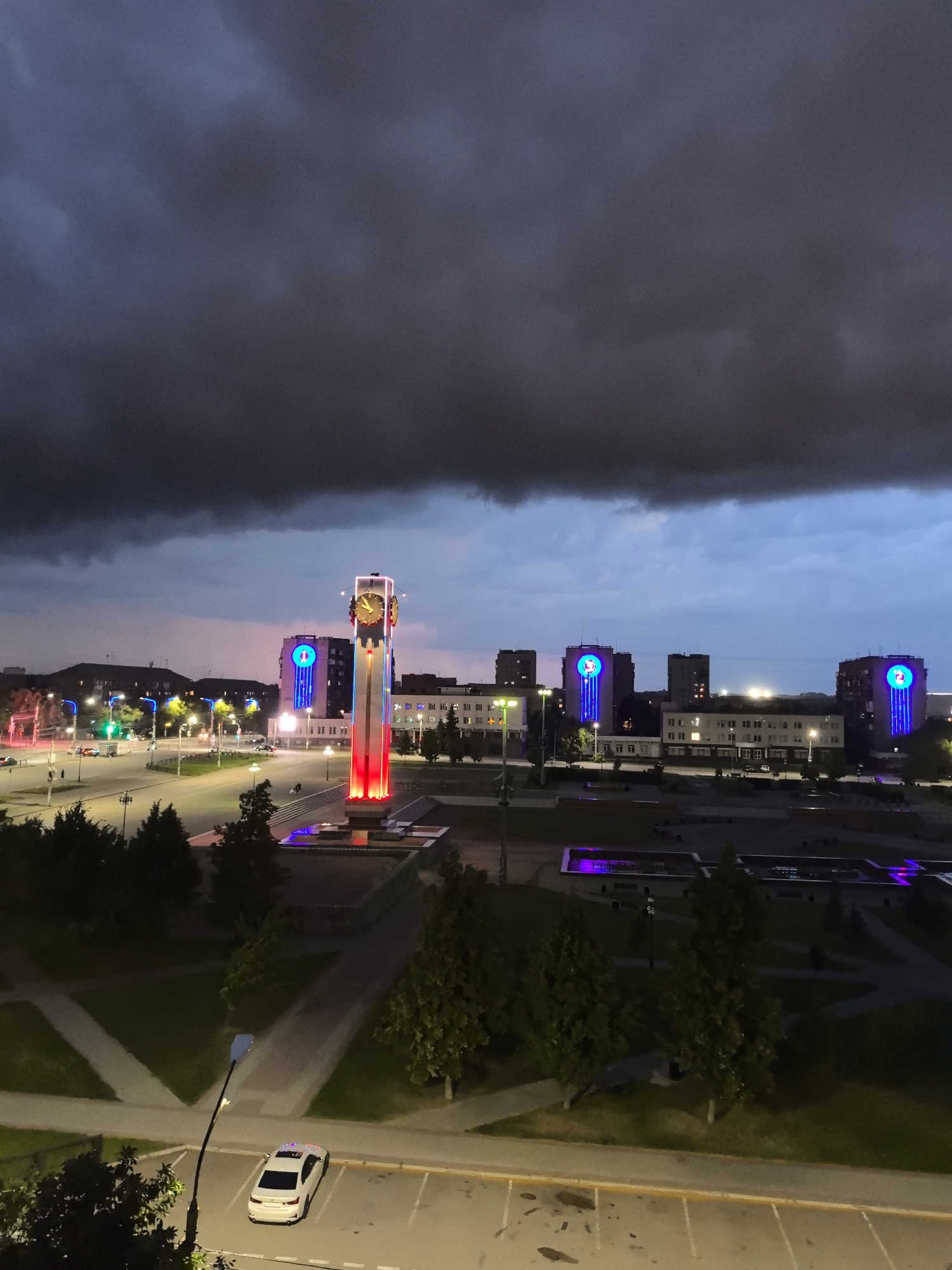 The image captures a nighttime view of the city of Magnitogorsk, Russia, likely from an elevated position. The foreground features a parking lot with a white sedan. A street lamp casts a glow on the asphalt. Further back, a park area with trees is visible, intersected by paved pathways. Dominating the midground is the iconic Magnitogorsk clock tower, illuminated with red and blue lights. Behind the tower, several multi-story buildings are present, some adorned with large, illuminated blue circular symbols. The sky above is dark and filled with dramatic, ominous clouds, suggesting an approaching storm. Streetlights and building lights create a vibrant urban scene against the darkening sky.