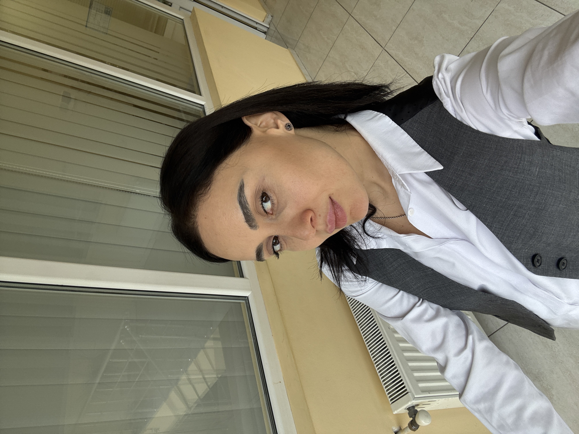 A woman with dark hair is facing the camera in a medium shot, wearing a white collared shirt and a gray vest. She has a neutral expression. The background consists of a yellow wall with a white window frame. The window has blinds. The woman is likely indoors or on a balcony.
The scene is not necessarily in Tbilisi, Georgia.