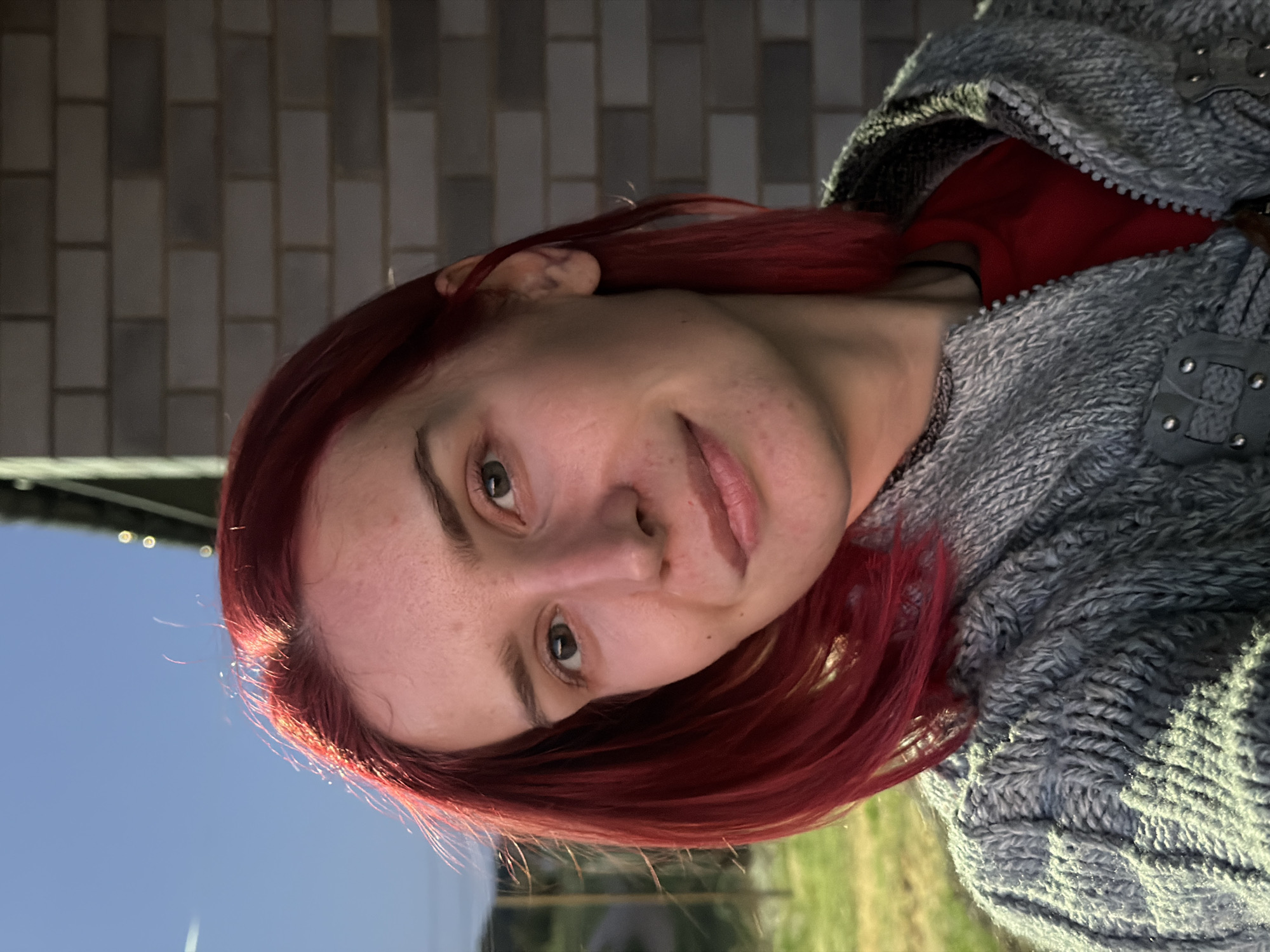 A medium close-up shot shows a young woman with vibrant red hair, facing the camera with a gentle smile. She is wearing a grey, textured knit jacket zipped over a red shirt. The background is a blurred mix of an outdoor setting with a brick wall to her right. The lighting suggests it is late afternoon or early evening, with the sun casting a warm glow on her face and hair. No specific location in Chaikovsky, Russia, can be identified from the image.