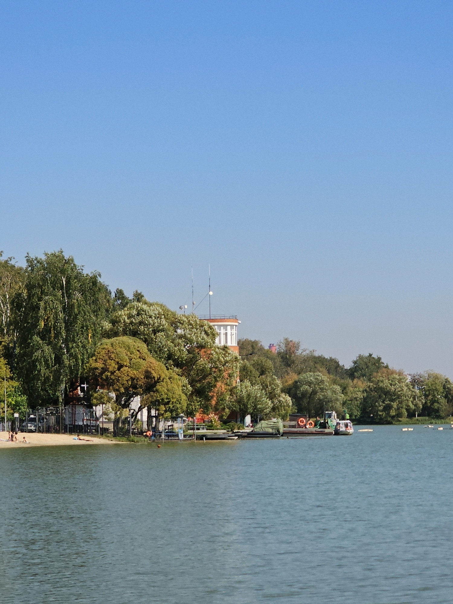 In Moscow, Russia, this image captures a serene waterscape with a tree-lined shore under a clear blue sky. The scene depicts a calm lake with a small sandy beach on its left bank, where a few individuals are seen relaxing. Some people are sunbathing on the sand, while others are wading or standing near the water's edge. A metal fence separates the beach area from the adjacent grassy embankment, which is densely populated with various trees, their foliage displaying a mix of green and autumnal hues.
On the right side of the image, a small floating platform or dock is visible, equipped with a few lifebuoys. A boat with a cabin is moored to this platform, and a few orange buoys are scattered across the water. In the background, a red and white building with windows stands amongst the trees, possibly a recreational facility or administrative building. The overall impression is one of a peaceful day in a park or leisure area, likely during late summer or early autumn.