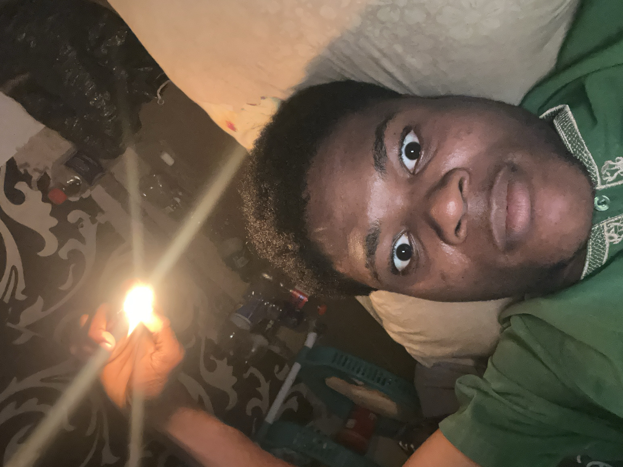 A person is lying down in bed, holding a lit lighter close to their face. The person has dark skin and is wearing a green shirt. The room appears to be dimly lit, with a patterned wallpaper in the background. The person's eyes are wide open, and they seem to be looking directly at the camera. There are some objects visible in the background, including bottles and a basket. The scene is likely a bedroom in a home in Ihiagwa, Nigeria, given the context provided.