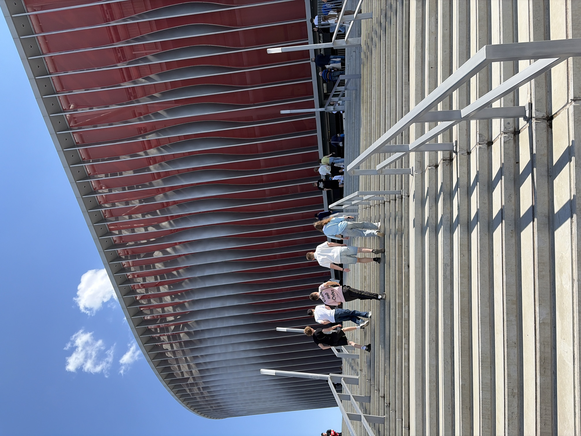 The image shows people walking up a set of concrete stairs adjacent to a large structure with a red and grey curved, slatted facade. The sky is blue with a few clouds. The location is likely the Dinamo National Olympic Stadium in Minsk, Belarus, based on the distinctive architectural features.