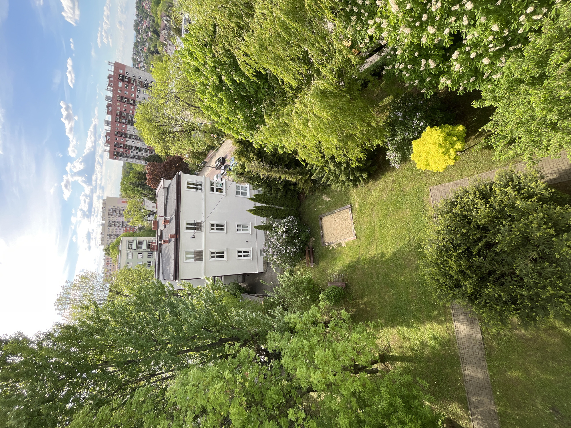 Here is a description of the image:
The image shows an aerial view of a courtyard in an urban setting. In the foreground, lush green trees dominate the bottom half of the frame. To the right, there is a brick walkway leading towards a green bush, with a patch of grassy area on either side of the walkway.
In the middle ground, a three-story white building with multiple windows is situated to the left. The building is adjacent to a parking area with a few cars. Adjacent to the building is a garden area with a sandbox and benches. The background includes other apartment buildings and rooftops visible against a blue sky with scattered clouds.
The scene appears to be taken on a bright, sunny day, with the sun's light creating shadows across the courtyard and buildings. The dominant colors are green, white, and blue.
