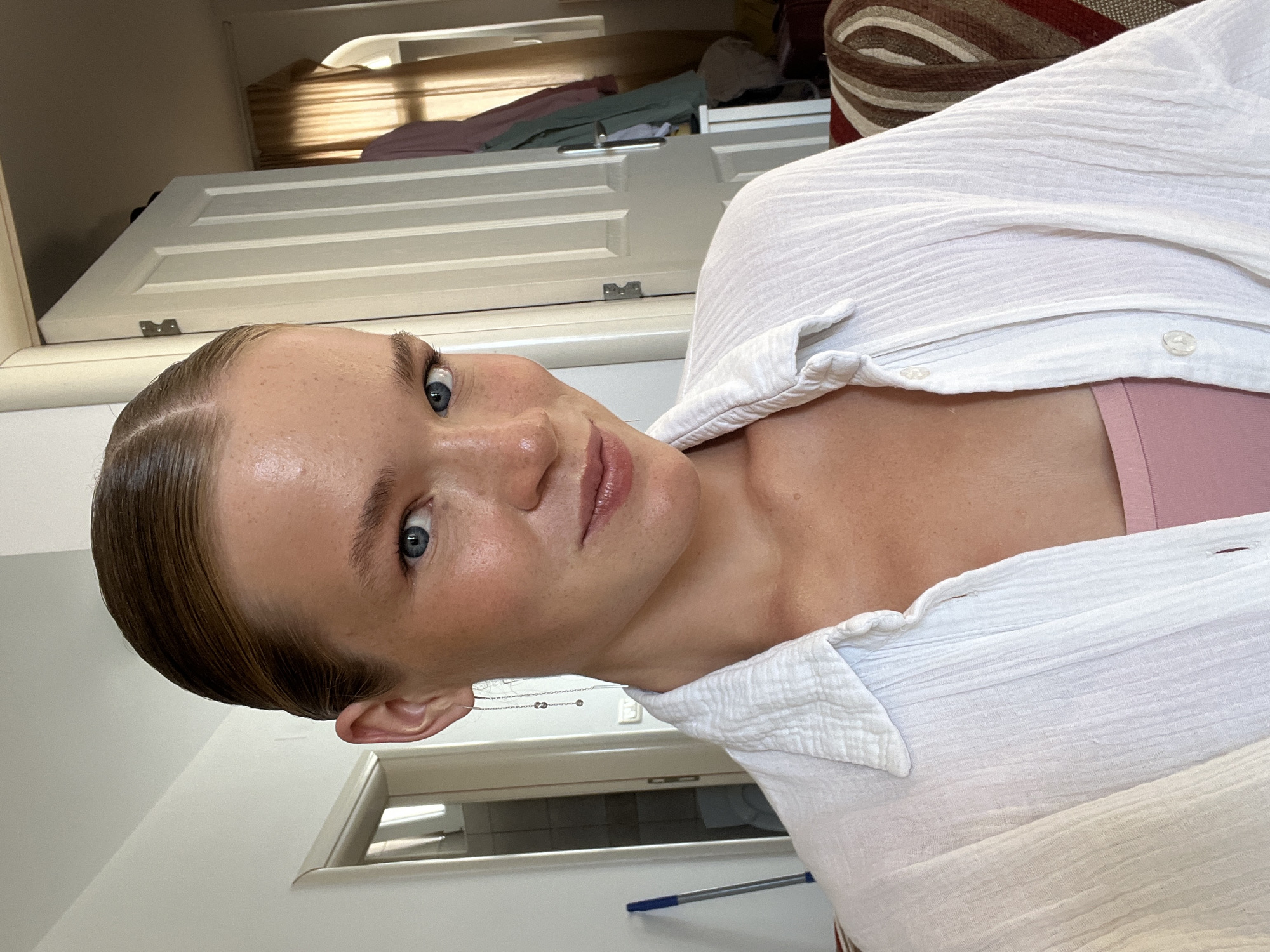 A young woman with blonde hair slicked back and blue eyes is captured in a selfie taken indoors. She is wearing a white open-collared shirt over a pink tank top. Her skin has a dewy glow, and she has freckles across her nose and cheeks. Delicate drop earrings with small stones hang from her ears. Behind her, a white door is visible, partially ajar, revealing a glimpse of a hallway or another room. To the right, another door with a wooden handle stands open, showing clothing draped on a rack and a glimpse of a window with a light-colored curtain. A mop with a blue handle leans against the wall to the left. The overall impression is a casual, personal moment captured in a domestic setting, possibly an apartment in Budva, Montenegro, given the architectural style of the doors and interior elements.