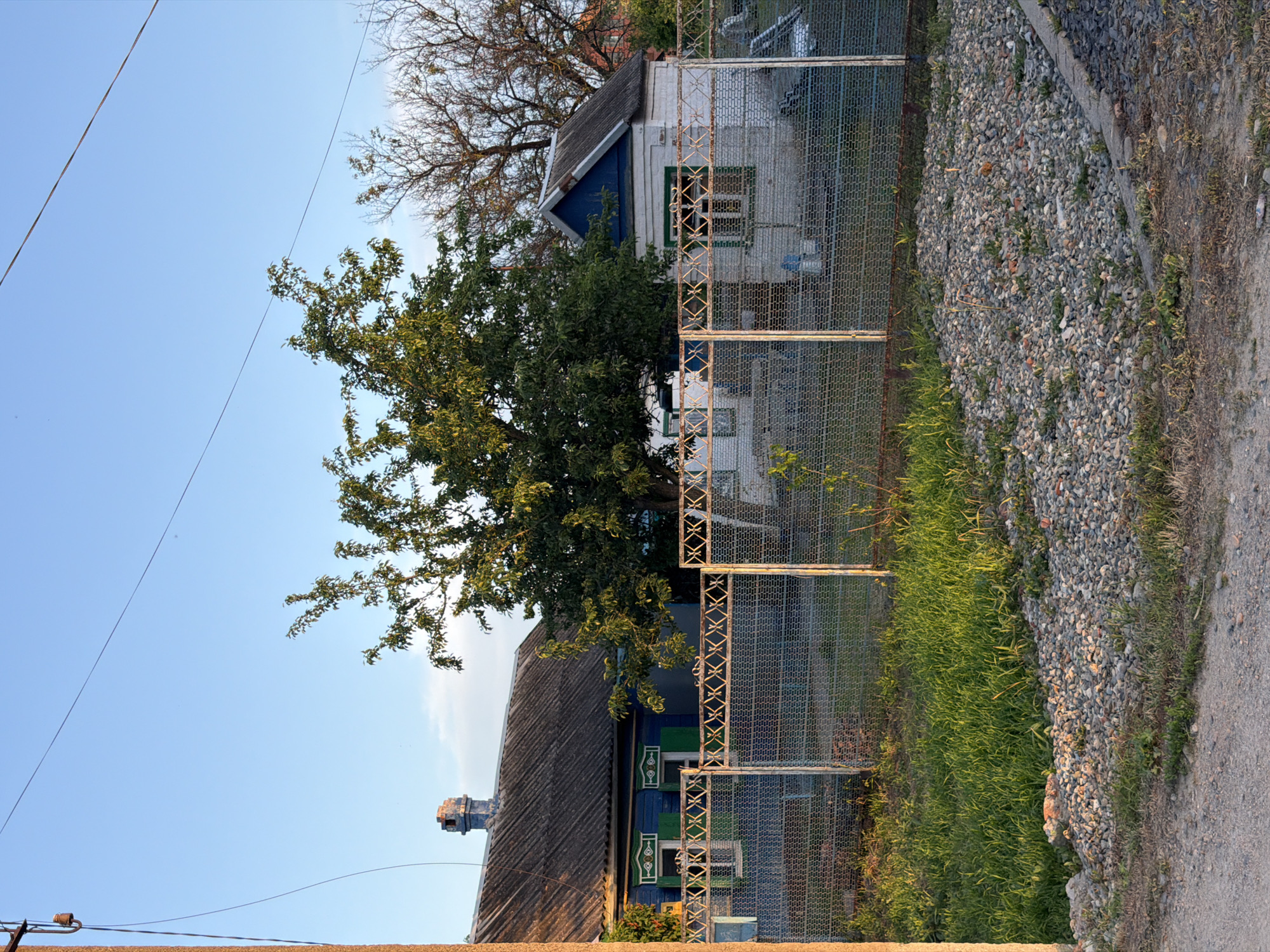 The image depicts a house and its surroundings in Leningradskaya, Russia. The house has a weathered appearance with blue trim and a gray roof. A tree obscures part of the house. A metal fence runs along the side of the house, separating it from a rocky area with some grass growing through it. A dirt road or path is visible next to the rocky area. Above, there's a clear blue sky.