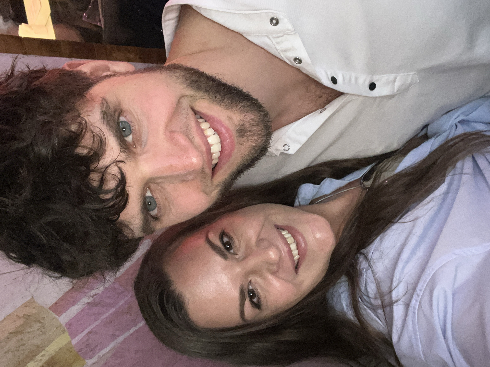Two people, a man and a woman, are posing for a photograph. They are lying down, likely on a surface with pink and white stripes.
The man is on the left. He has curly dark brown hair, blue eyes, and a short beard. He is wearing a white button-down shirt.
The woman is on the right. She has long dark brown hair and brown eyes. She is wearing a light blue button-down shirt.
Both individuals are smiling.
No specific location is identifiable in this image.