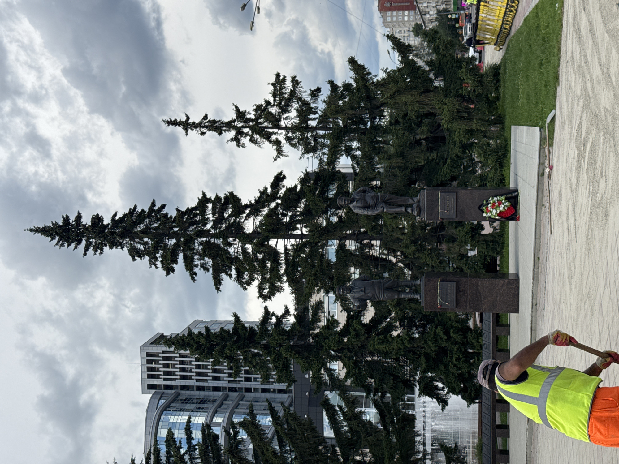 In Yekaterinburg, Russia, a worker in a high-visibility vest and orange pants is using a shovel to move material on a paved area in front of a monument. The monument features two bronze statues of men on granite pedestals, one with binoculars and the other with his hand in his pocket. A wreath of red and white flowers sits at the base of one pedestal. Behind the monument, tall evergreen trees frame modern buildings, including one with a distinctive curved glass facade. The sky is overcast with heavy clouds, suggesting impending rain. In the background, a fountain is visible, and further down the street, traffic and yellow barriers indicate ongoing construction or maintenance.