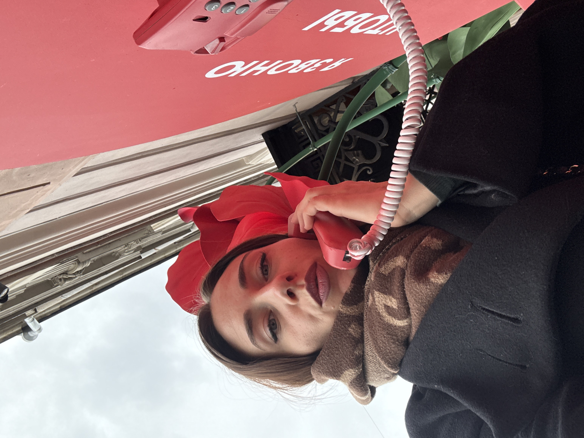 A woman is seen at eye level in the city of Saint Petersburg, Russia, holding a red telephone receiver to her left ear with her left hand. She is wearing a brown patterned scarf and a black coat. She has brown hair and is wearing makeup. She is standing underneath a red kiosk with white writing, which reads "170561 ЯЗВОНКИ". Above the kiosk, there is a green metal structure. Behind the kiosk, there is a building with a white exterior. The sky is overcast.