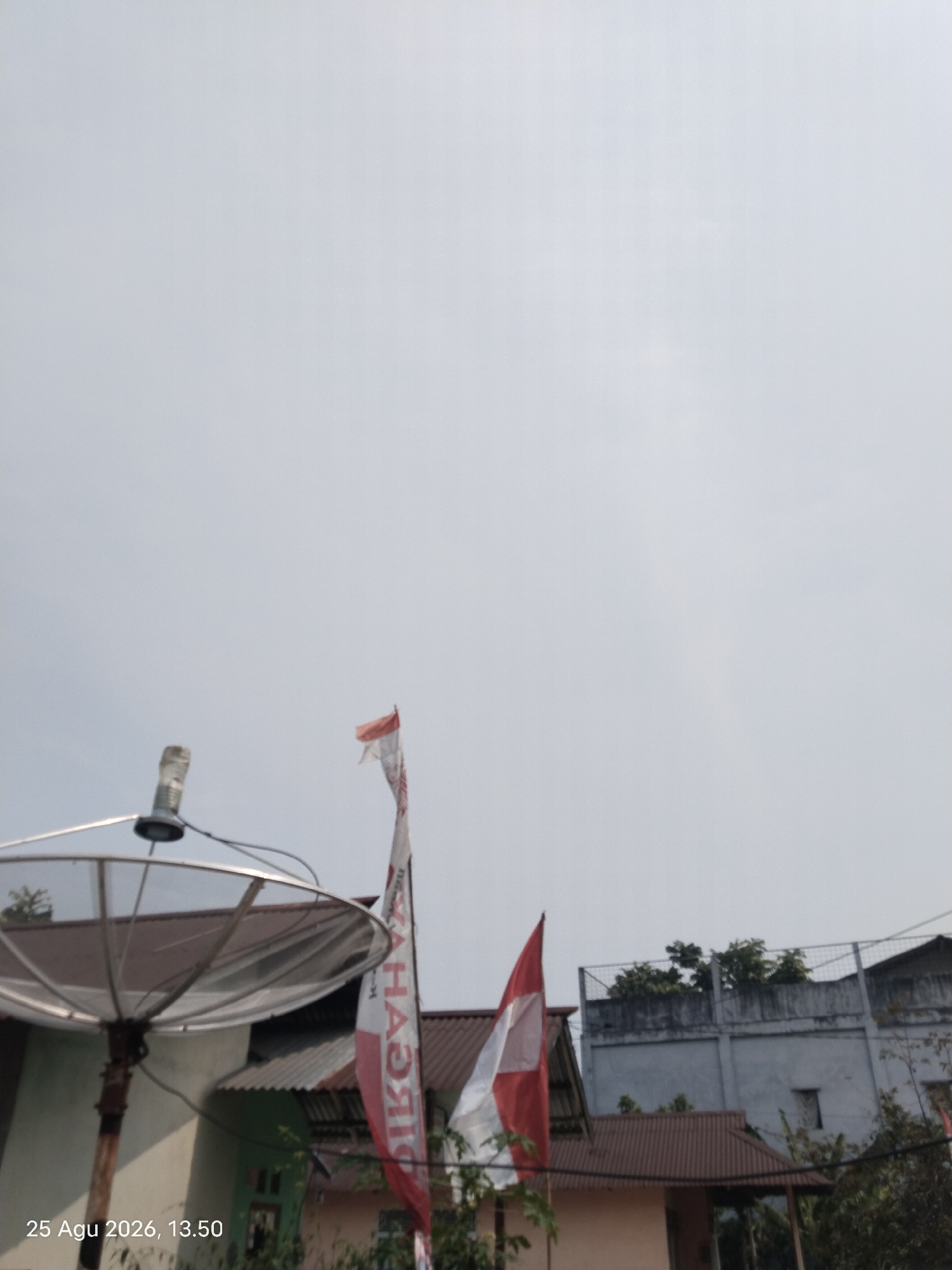 The image shows a cloudy sky with a satellite dish in the foreground on the left. Two Indonesian flags are visible in the middle of the frame, one slightly behind the other. The flags are red and white, with text visible on the taller, further flag. Buildings with brown and light-colored roofs are present behind the flags, and a taller, grey building with a fence on top is in the background on the right. Trees are visible around the buildings. The date and time stamp "25 Agu 2026, 13.50" is present in the bottom left corner of the image. The scene appears to be an outdoor setting in a residential or urban area, likely in Singkawang, Indonesia, given the presence of Indonesian flags and the date stamp. There are no people visible in the image.