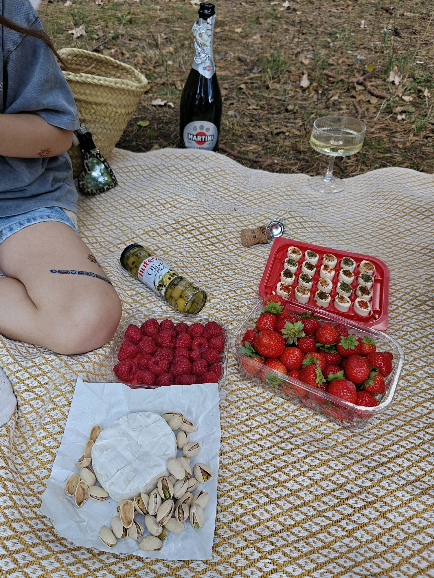 A person is having a picnic on a checkered blanket in Piaski, Poland. The picnic includes a bottle of Martini Asti champagne, a glass of champagne, a basket, a jar of olives, a container of strawberries, a container of raspberries, a wheel of brie cheese surrounded by pistachios, and a tray of small appetizers. The person is wearing a grey t-shirt and denim shorts. The ground is covered in pine needles and leaves.
