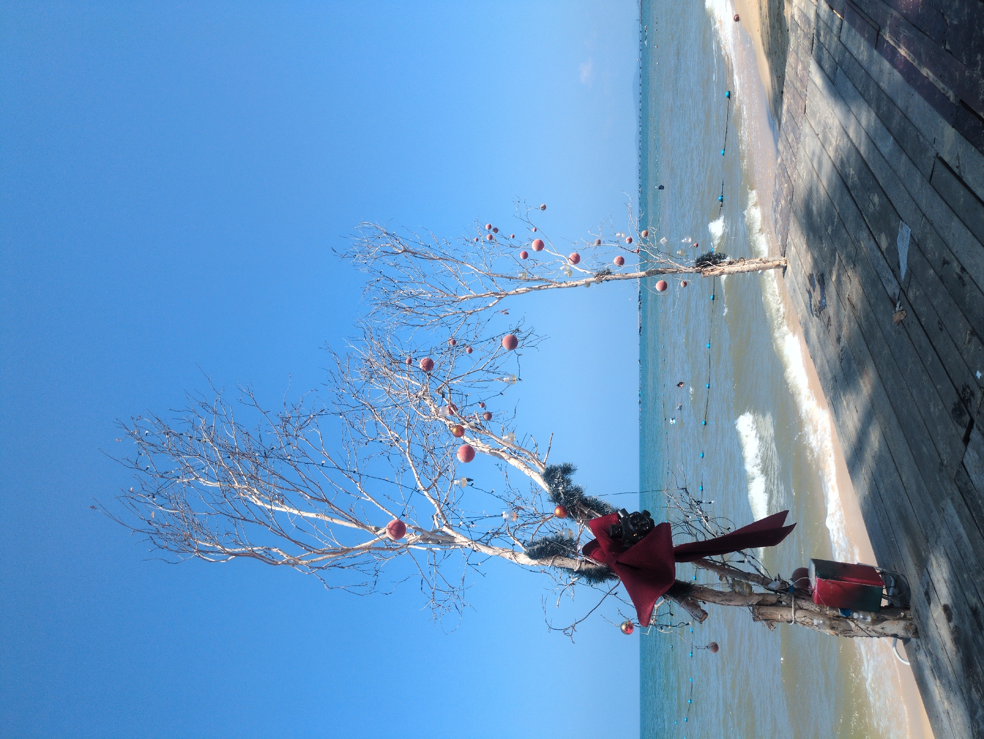 The image shows a makeshift Christmas tree made of bare branches and red ornaments, standing on a beach in Phú Quốc, Vietnam. The tree is decorated with a large red bow and tinsel. Another similar tree stands farther away on the beach. The background consists of a blue sky and the ocean, with gentle waves lapping the shore. A wooden boardwalk or platform is visible on the right side of the frame. The scene appears to be a festive holiday display set up on the beach.