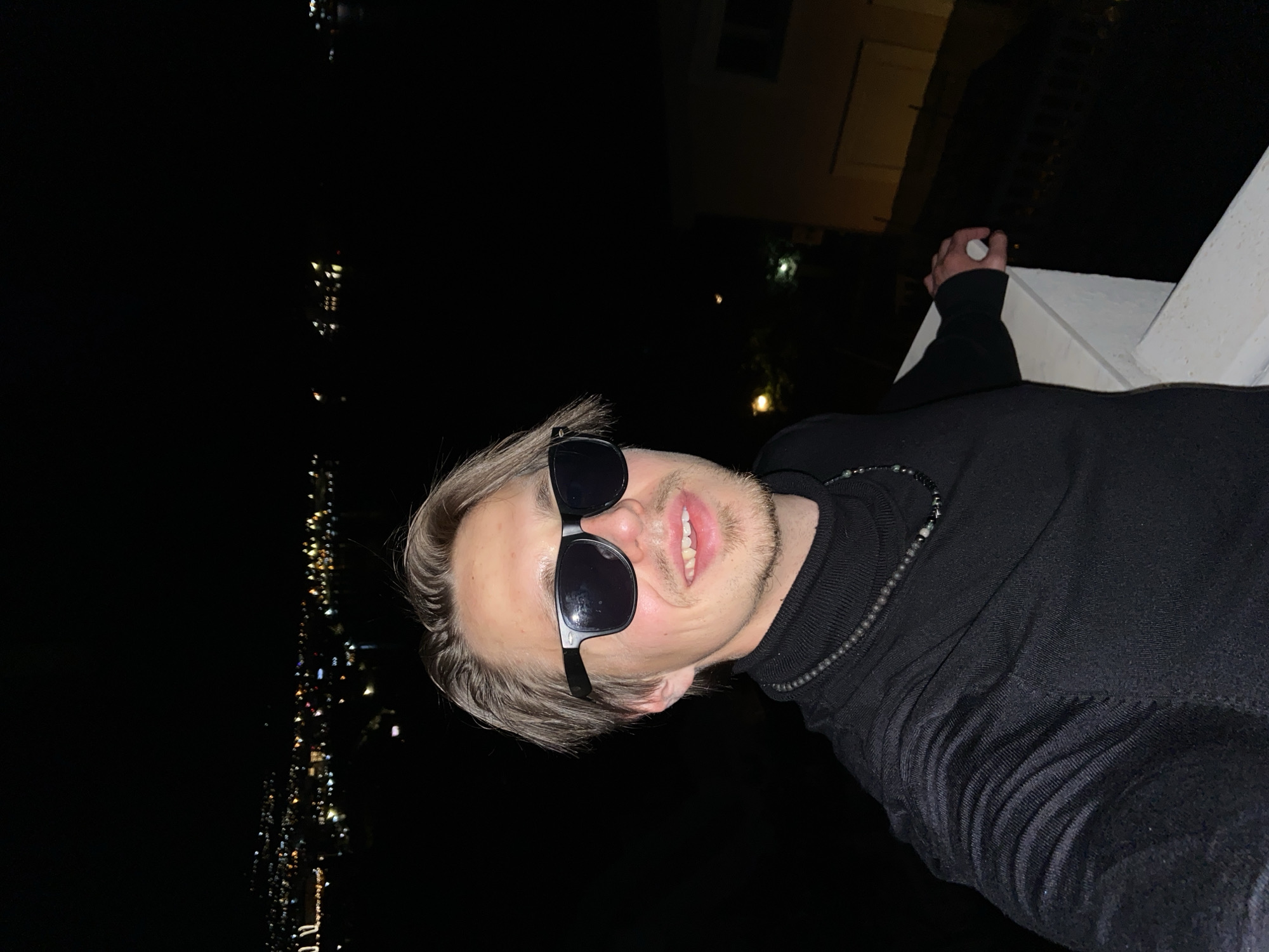 A person with blond hair and dark sunglasses is the subject of the selfie-style image. They wear a black turtleneck sweater and a beaded necklace. The person is leaning against a white ledge or railing. In the background, there's a night cityscape, with multiple light sources visible. A building is visible in the upper right quadrant of the image. The location may be Šušanj, Montenegro, based on the context. The photo appears to be taken at night.