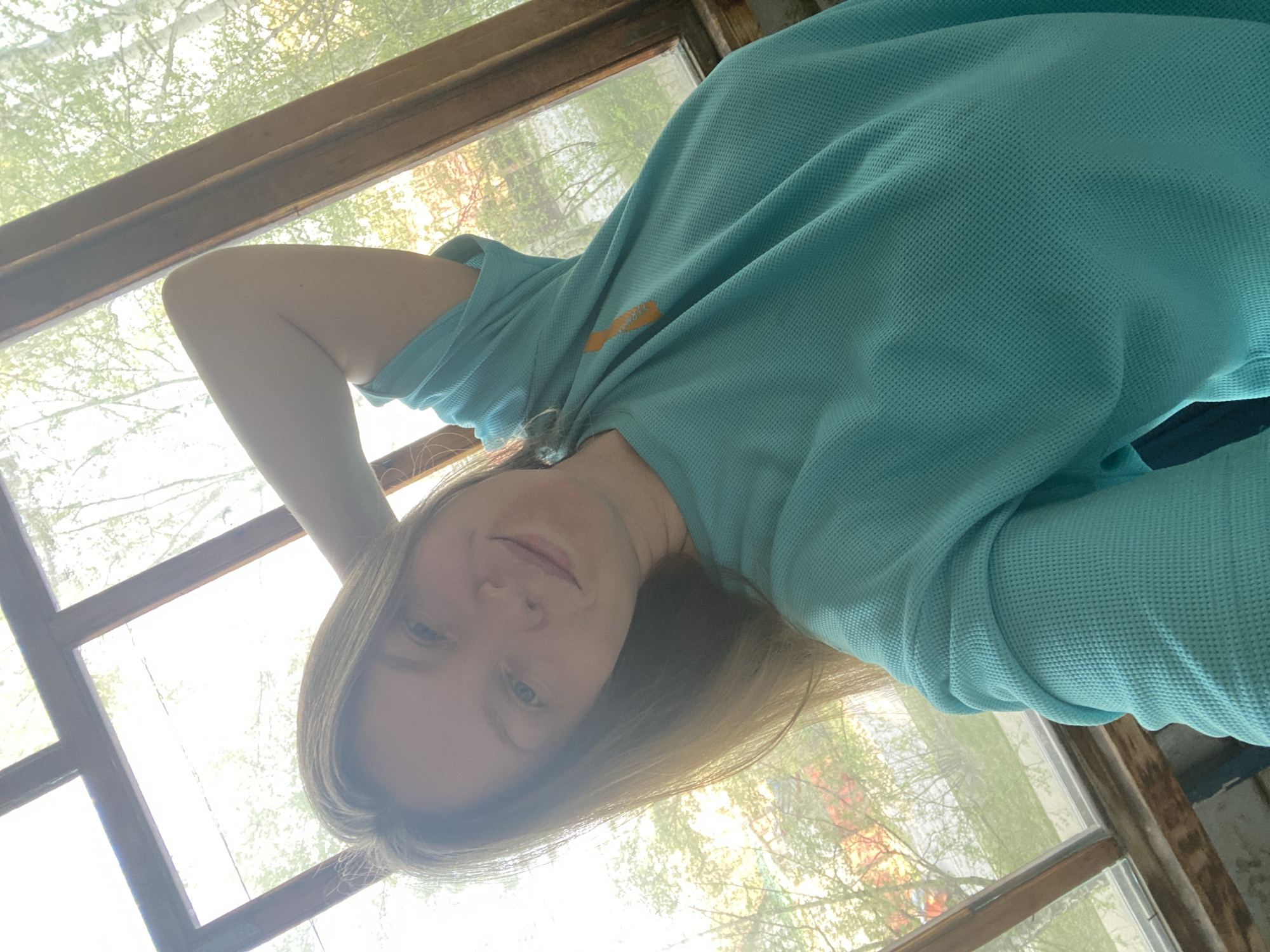 A woman with light hair and blue eyes is taking a selfie indoors, looking down at the camera. She is wearing a turquoise shirt with a small orange logo on the left shoulder. Her left arm is raised above her head. The background consists of a window frame, with trees visible through the glass. The lighting is bright, potentially from the sun shining through the window. There is no location in the city of Noviy, Russia.