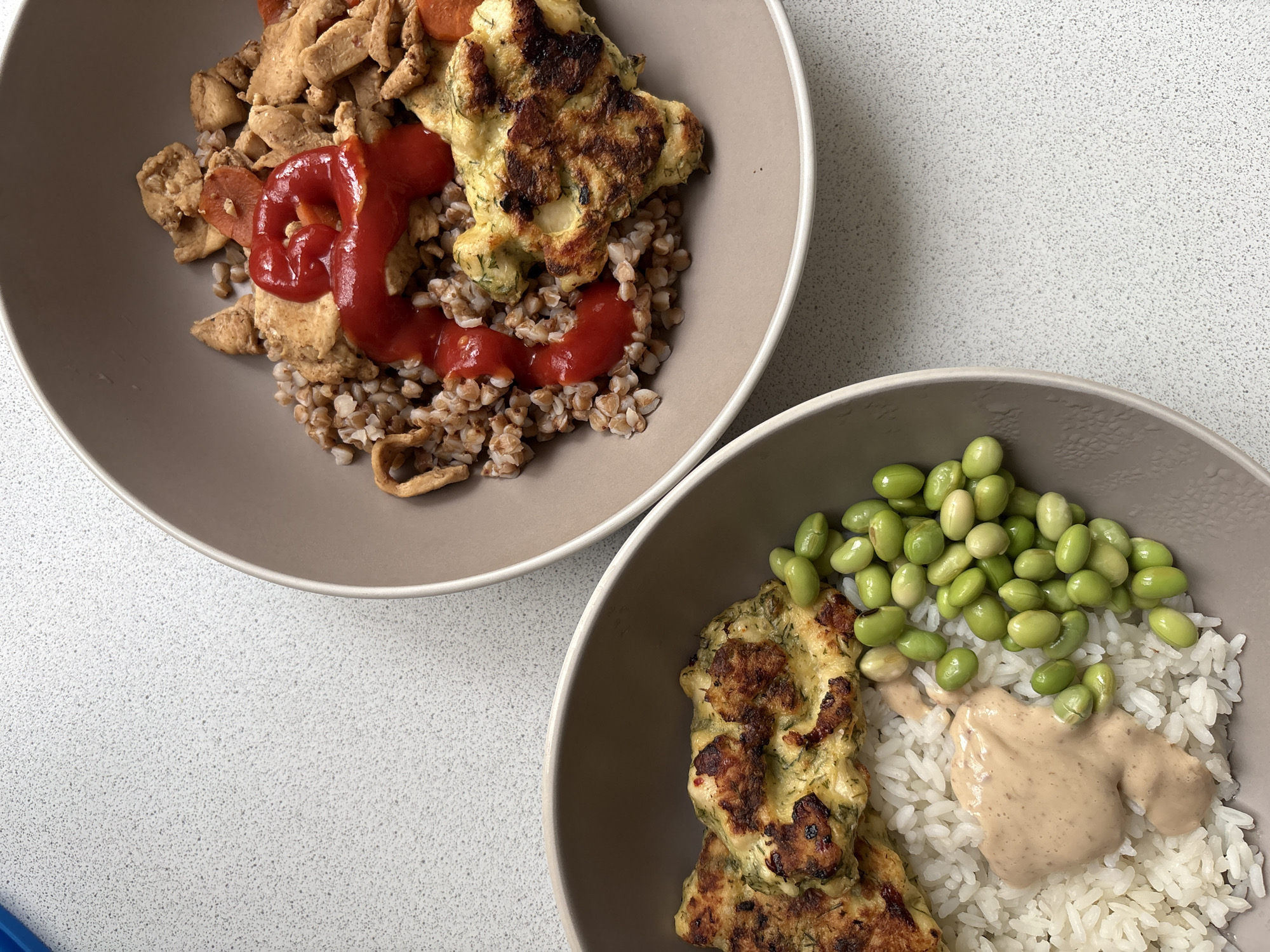 Two light beige bowls of food are displayed on a light gray, speckled countertop.
The top bowl contains a serving of buckwheat, topped with diced, cooked chicken or meat, sliced orange carrots, and a generous dollop of bright red ketchup or similar sauce. Alongside these are two golden-brown, pan-fried patties or fritters, appearing to contain green herbs like dill.
The bottom bowl features white rice as its base, adorned with a scattering of vibrant green edamame beans and a swirl of creamy, light brown sauce. Similar to the top bowl, it also holds two golden-brown patties or fritters, identical in appearance to those in the first bowl, suggesting they are from the same batch.
The scene depicts two distinct, home-cooked meals, possibly for two individuals or offering variety to one person. No people are visible. There is no visible text. The lighting suggests an indoor setting during the daytime. Although the location context is Saint Petersburg, Russia, there are no specific visual cues within the image to confirm this.